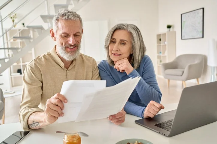 An older couple sitting at table, looking over documents and laptop, smiling at home