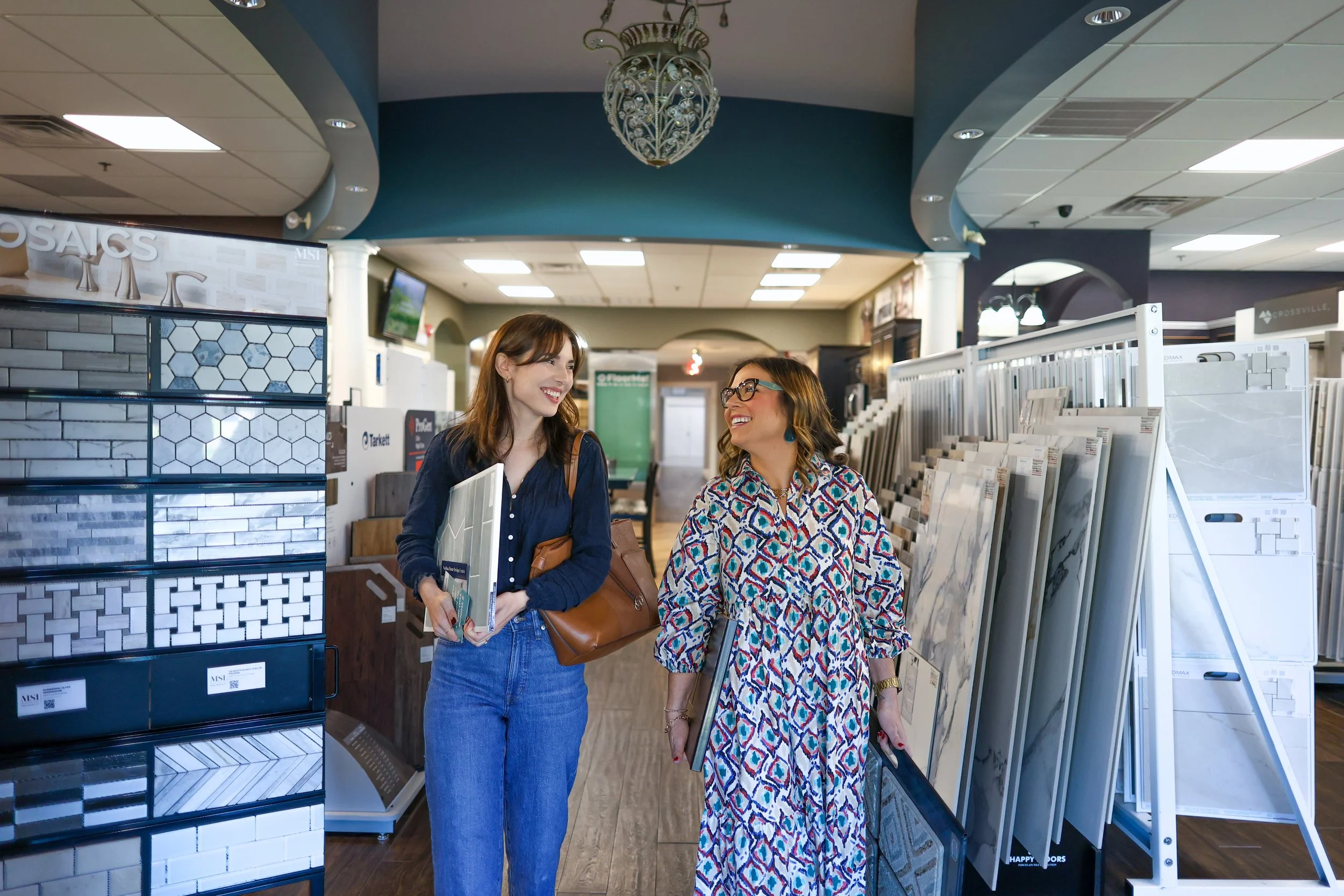 Two women smiling and walking through a tile showroom, surrounded by displays of tiles and flooring samples.