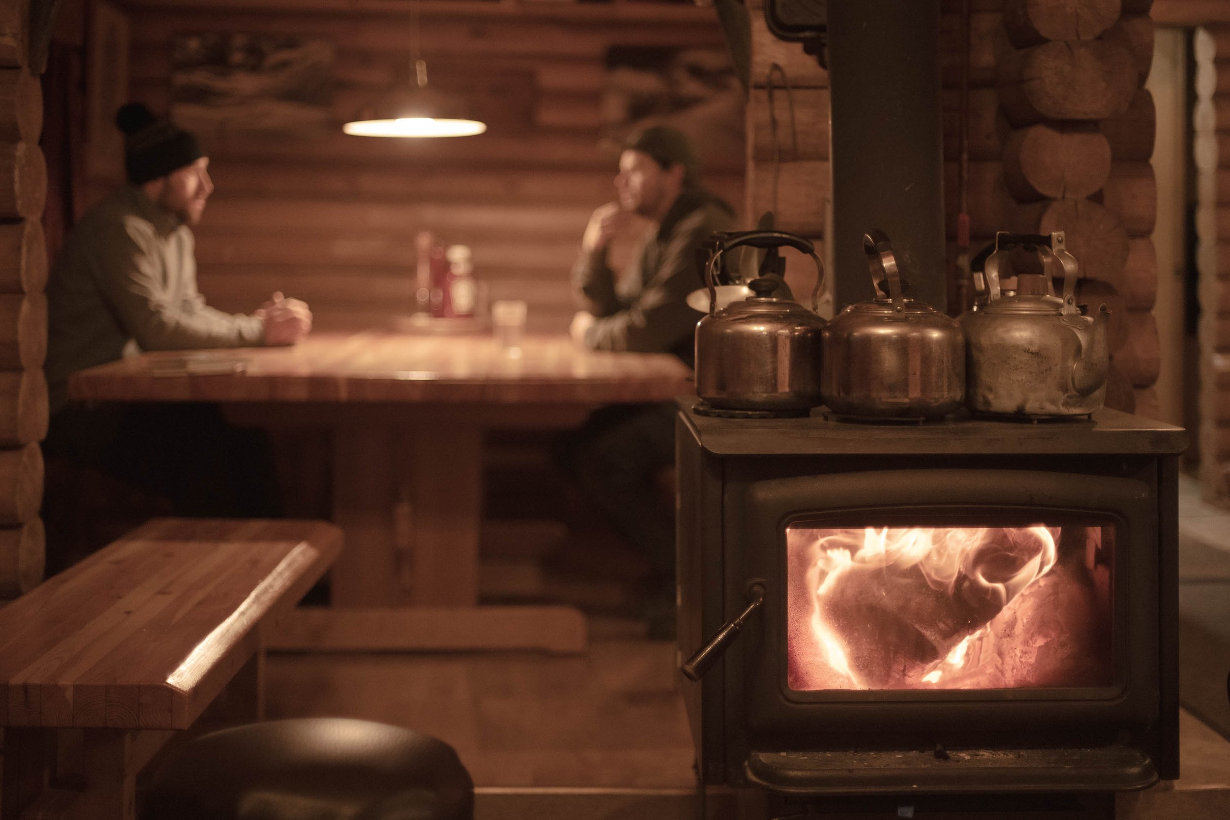 Two people sitting at a wooden table conversing inside a cozy log cabin-style room, with a stove warming the space and four kettles on top of it. toursim photography in banff alberta