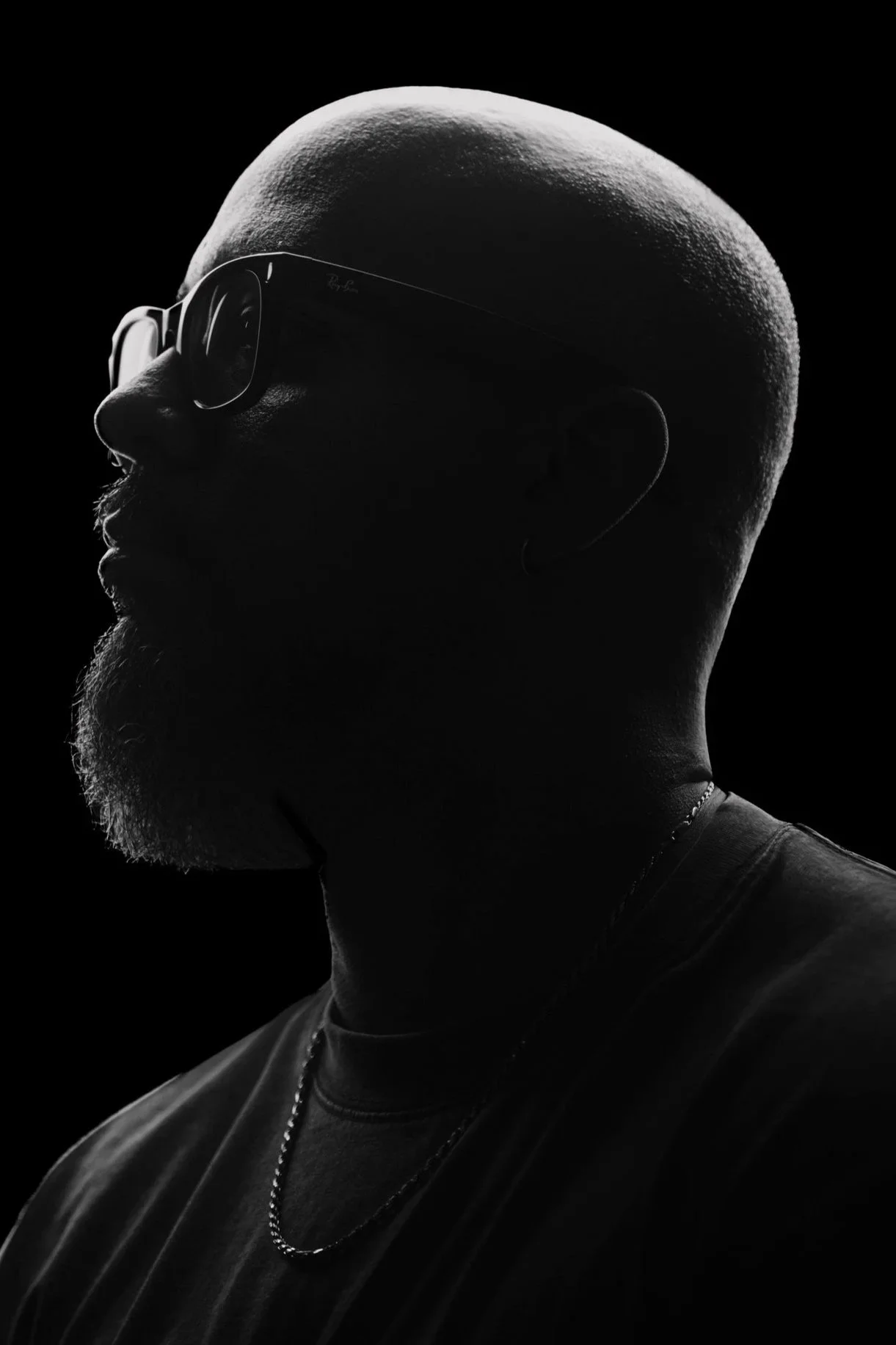 Black and white silhouette of a man wearing sunglasses, with a shaved head, beard, and a chain necklace, looking to the side against a black background.