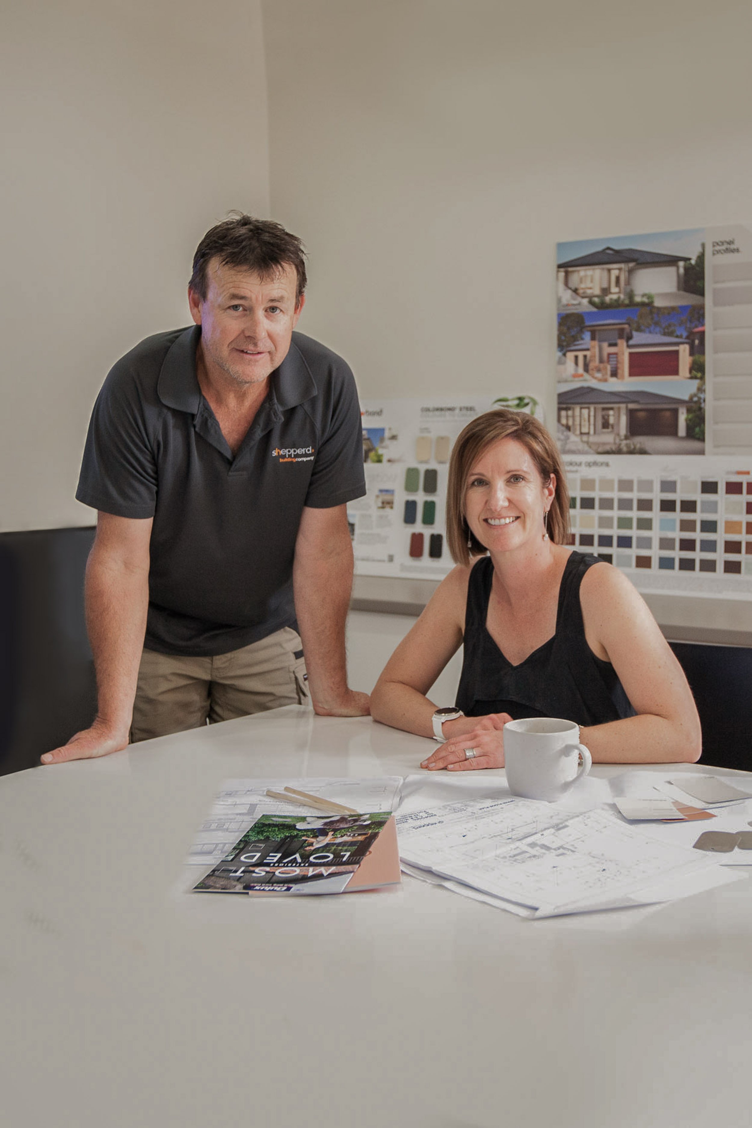 A man and a woman are sitting at a white table in an office or meeting room. The man is standing and leaning on the table, wearing a dark gray polo shirt with an orange logo, and the woman is seated, wearing a black sleeveless top. She is smiling, with a white mug in front of her, and there are papers and magazines on the table. Behind them are posters and color samples related to home design.
