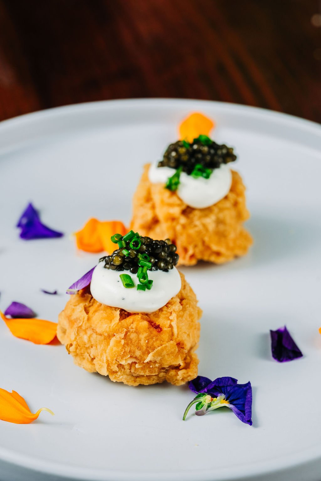 Two fried chicken bites topped with sour cream, chopped green onions, and caviar, garnished with edible flower petals on a white plate.