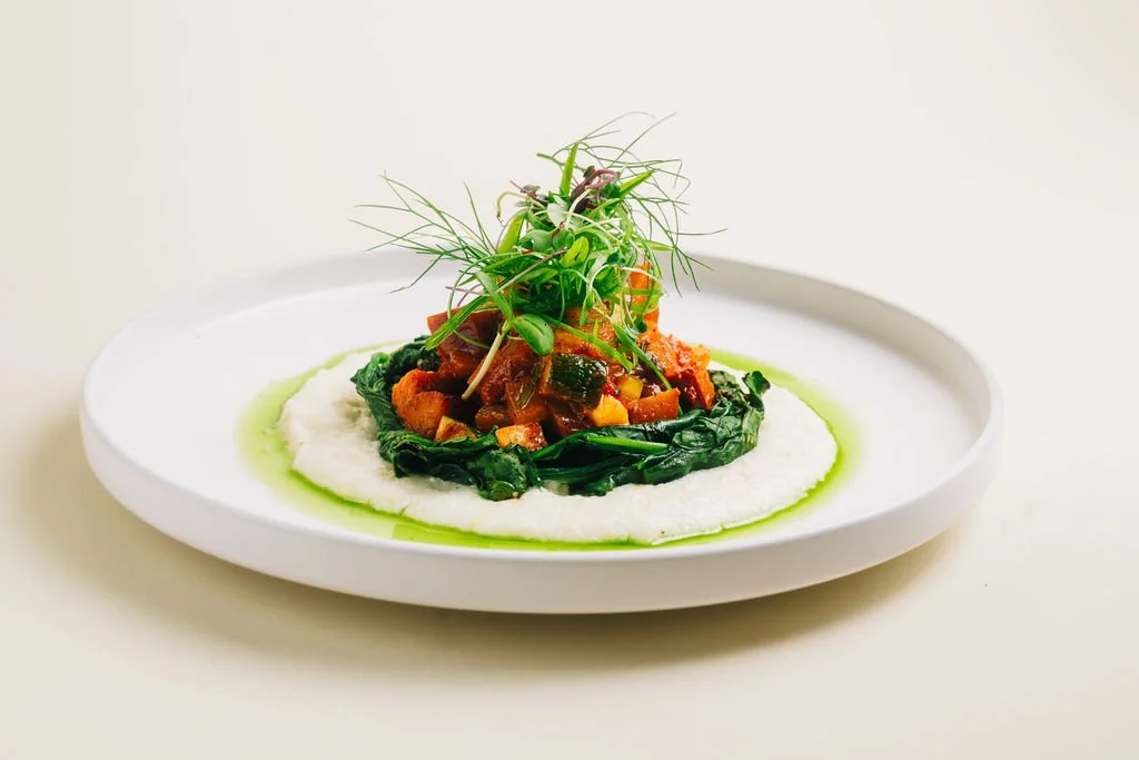 Gourmet dish with roasted vegetables on a bed of greens, topped with microgreens, served on a white plate with green sauce.