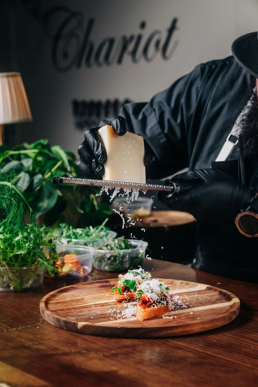 Person wearing black gloves and a black shirt grating cheese over a plate of food on a wooden table, surrounded by green plants and containers of salad.