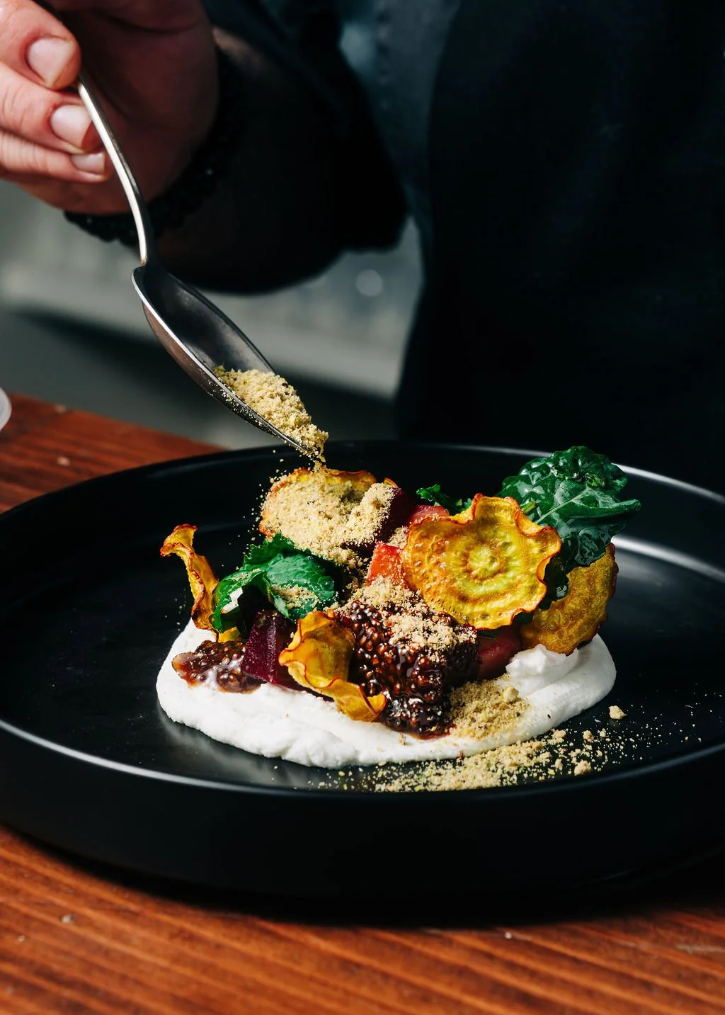 Chef sprinkling yellow powdered seasoning onto a plated gourmet dish with roasted vegetables and sauces.