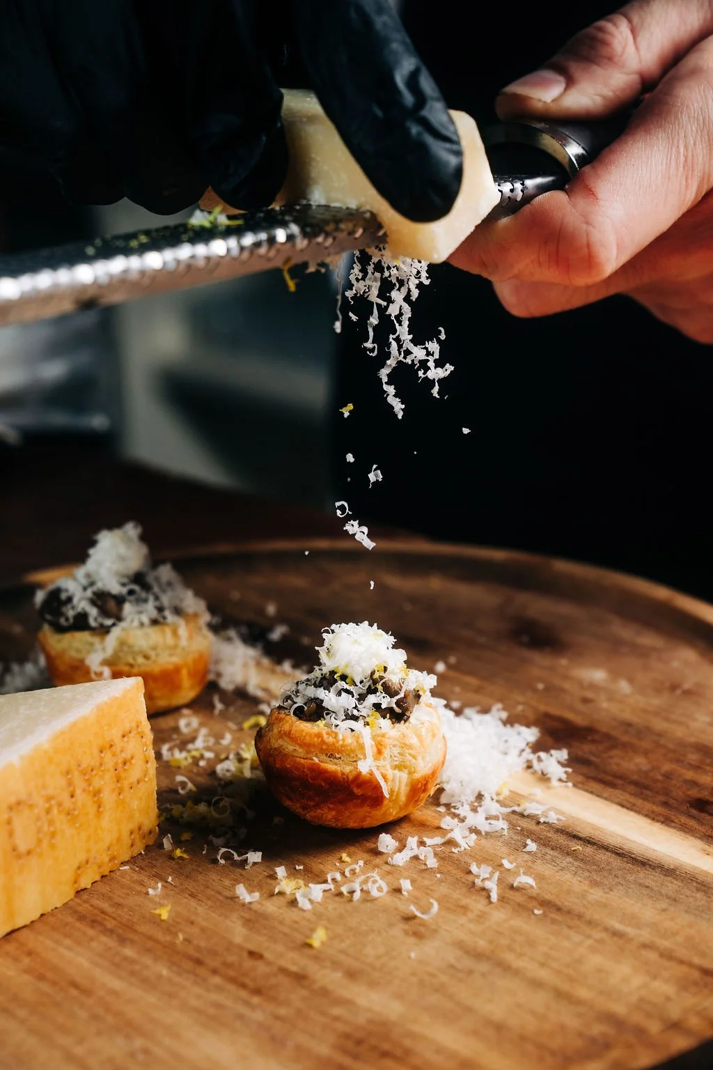 Close-up of a person grating cheese onto small savory pastries with dark filling on a wooden cutting board.