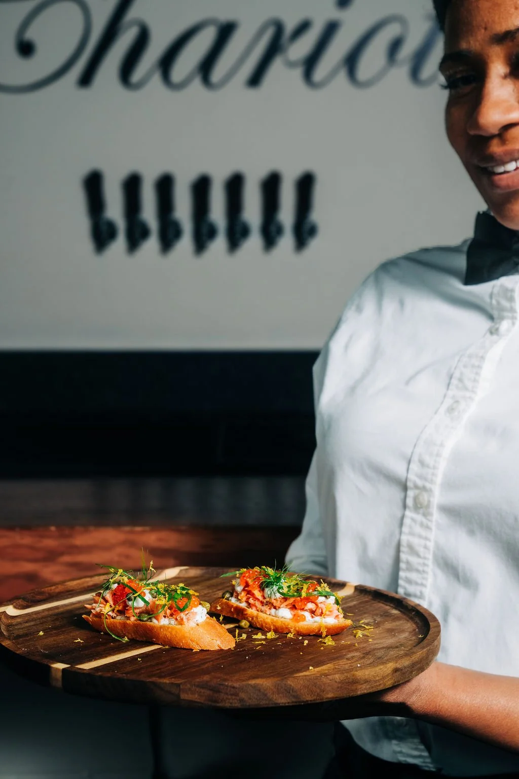 A person holding a wooden serving platter with two open-faced sandwiches topped with herbs, vegetables, and seafood, in a restaurant or dining setting.