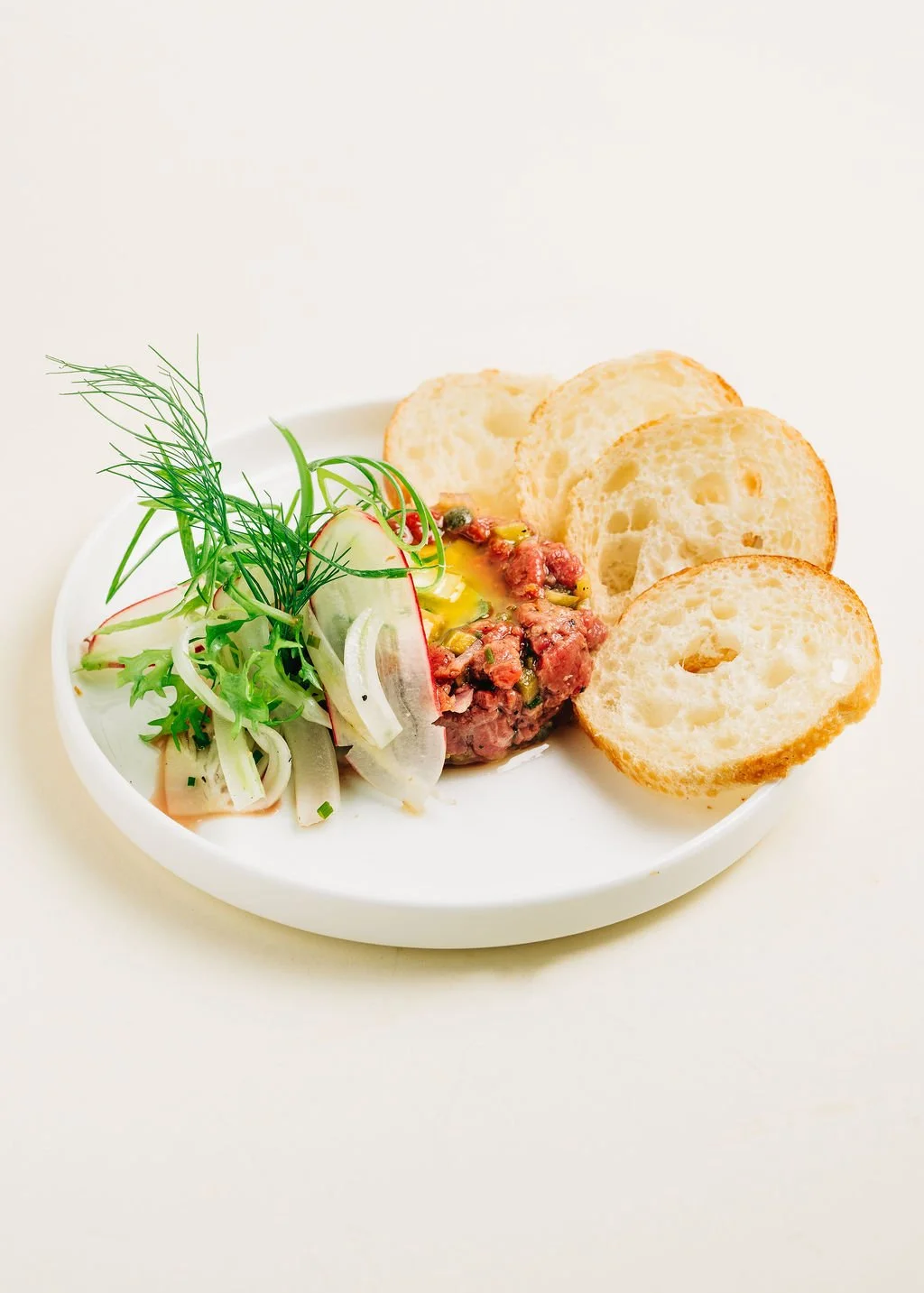Appetizer plate with sliced baguette, fresh greens, and steak tartare garnished with herbs.