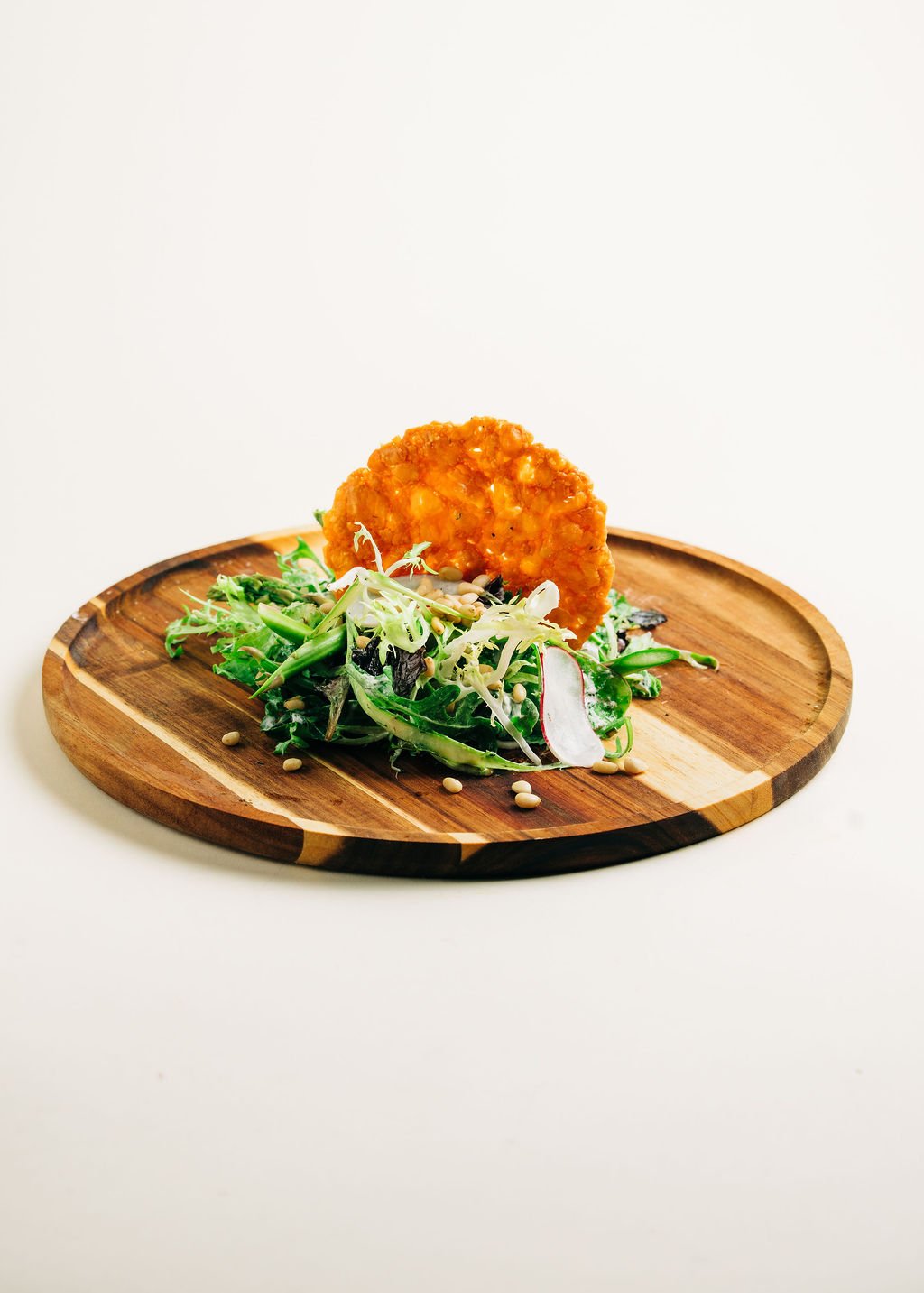 Salad with mixed greens, radish slices, sunflower seeds, and a crispy fried snack on a round wooden plate.