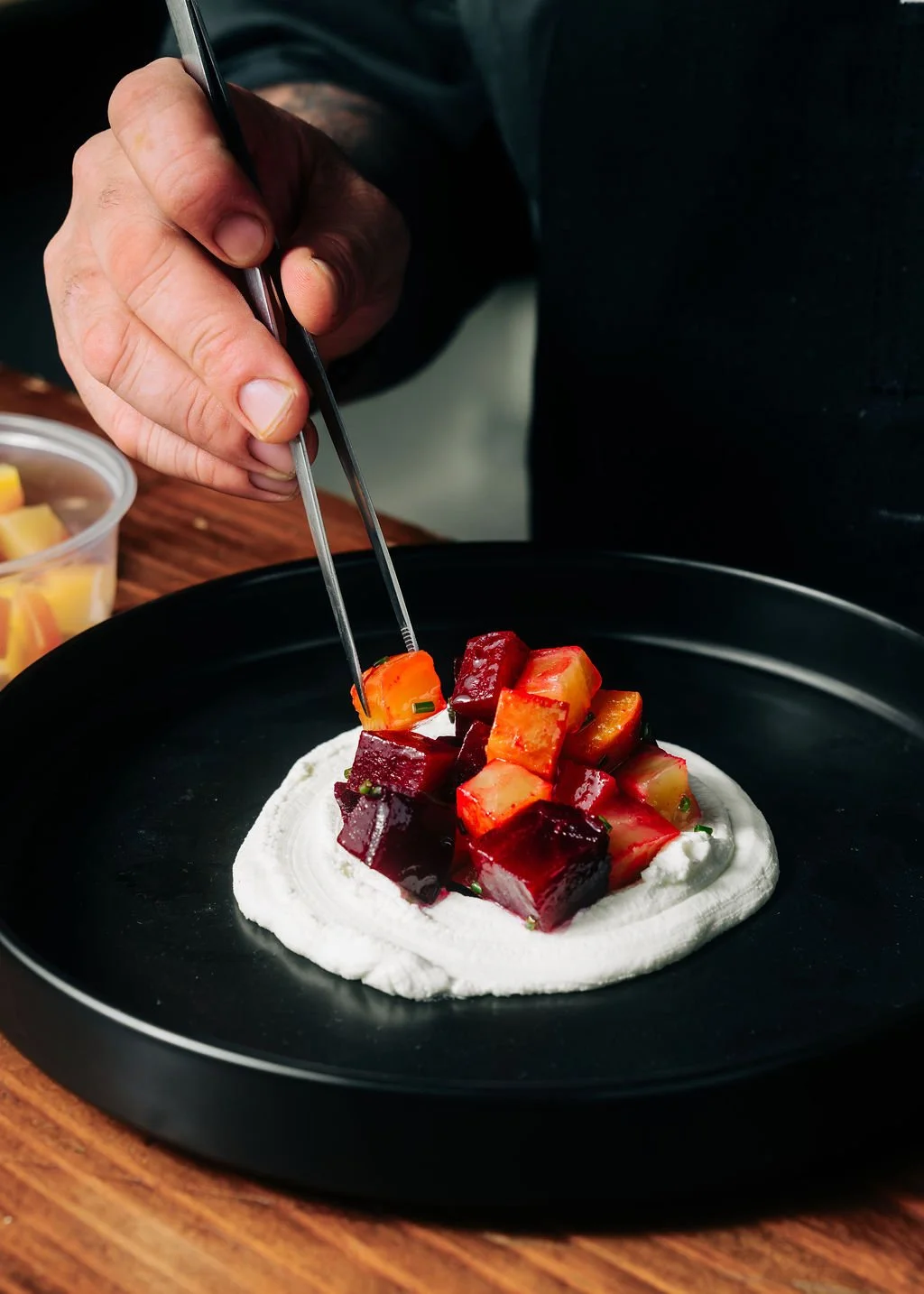 A chef garnishing a dish with diced colorful vegetables on a creamy base on a black plate.