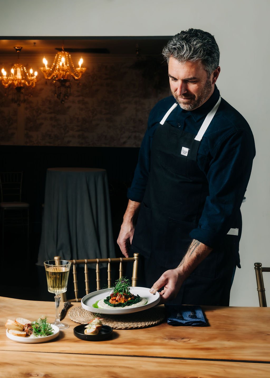 A chef in a black uniform and apron setting a plate of gourmet food on a wooden table, with a glass of white wine and a small plate of appetizers nearby. Elegant chandeliers hang in the background.