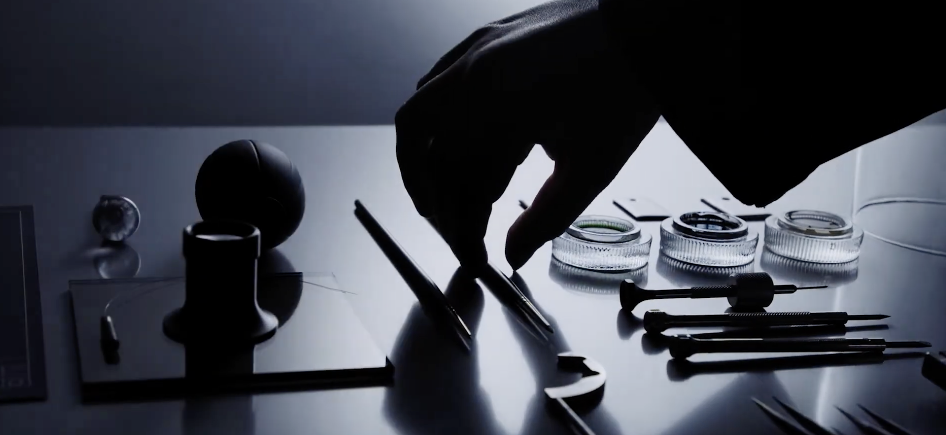 A silhouetted hand reaching for small glass petri dishes on a lab table, with various laboratory tools like syringes and scalpel nearby, illuminated by backlighting.