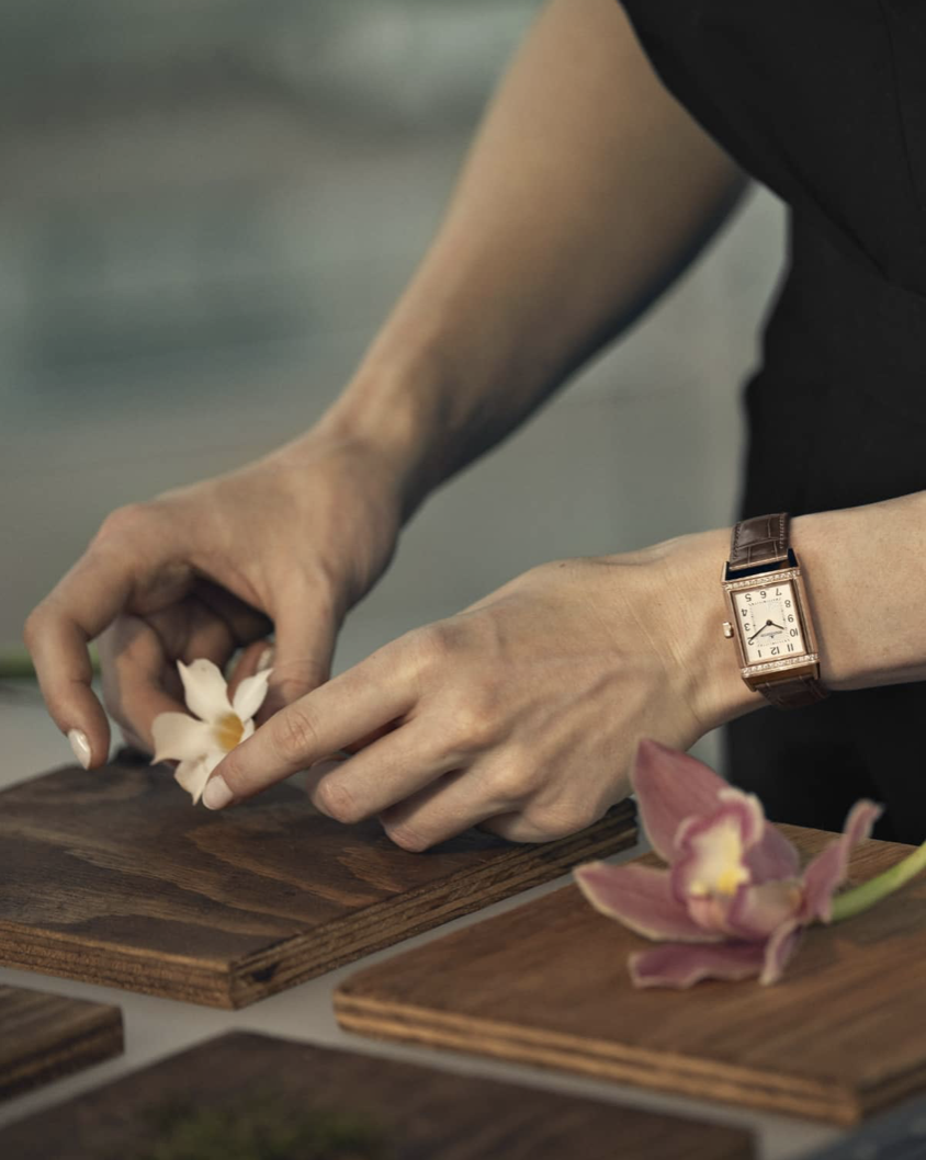 Hands arranging a white flower on a wooden surface, with a pink orchid in the foreground, wearing a vintage-style watch with a brown leather strap.