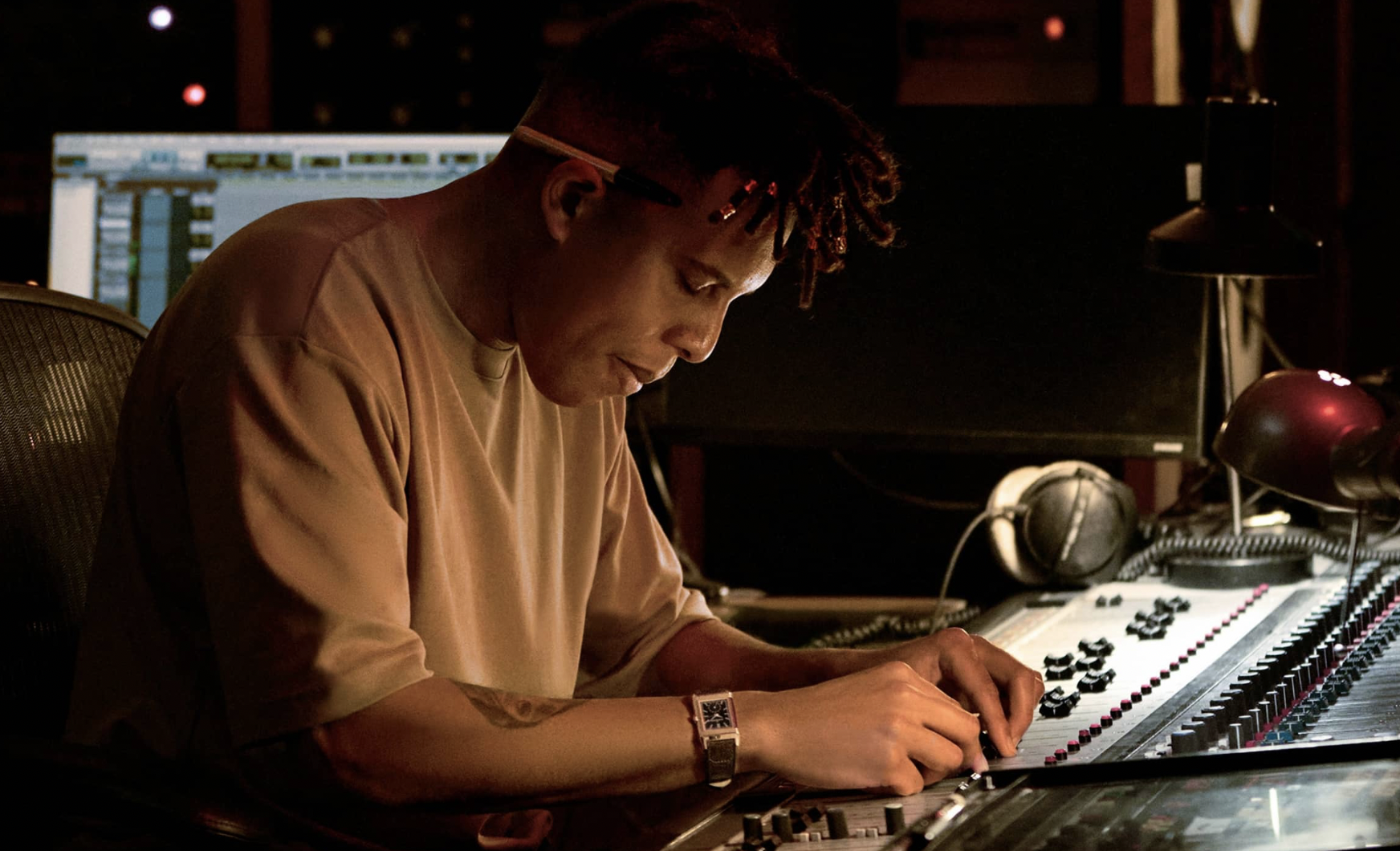 A person with glasses and a beige shirt, working at a sound mixing console with a computer screen in the background, in a recording studio.