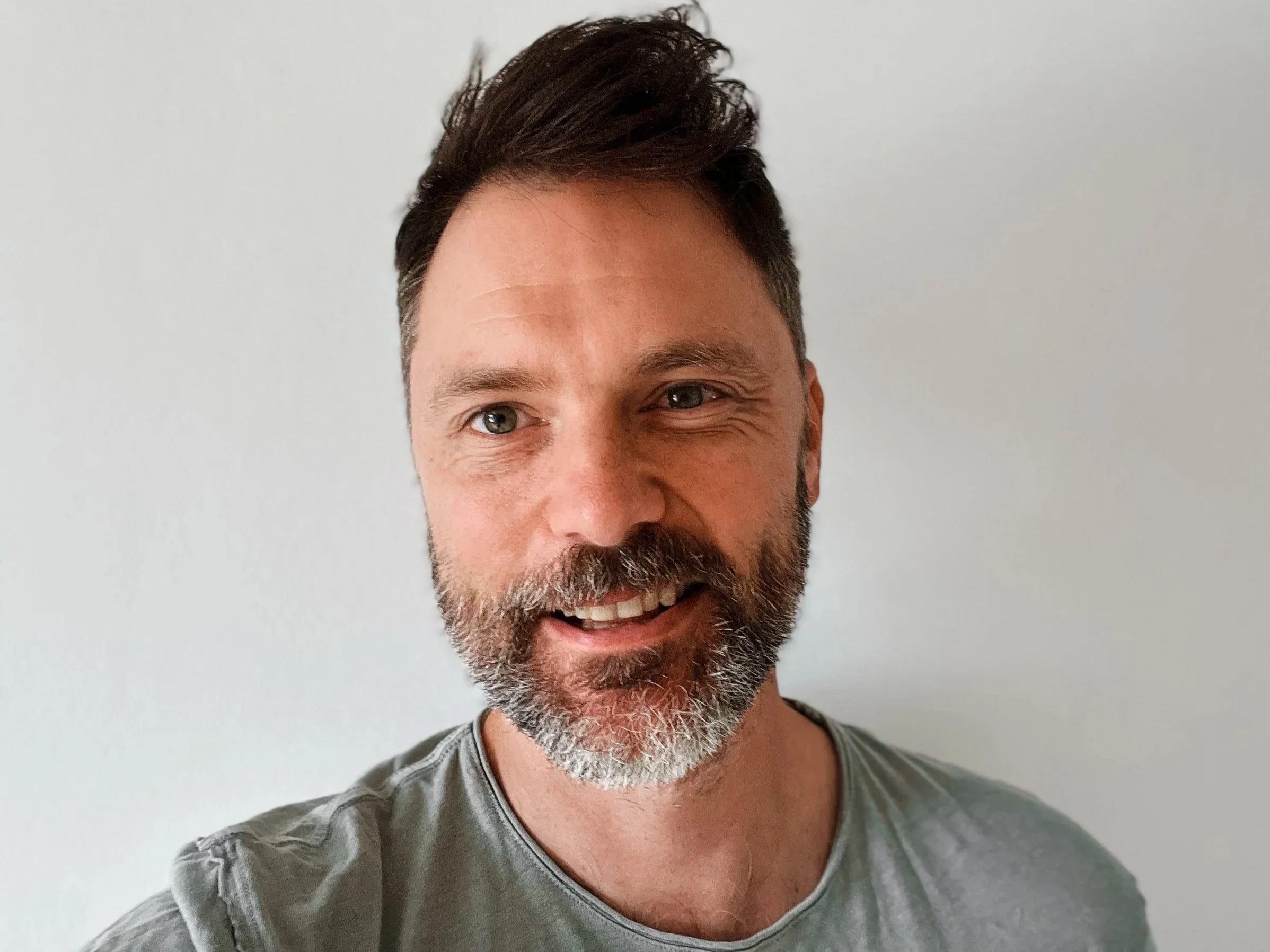 A smiling man with a gray beard and short dark hair, wearing a gray shirt, standing against a plain white background.
