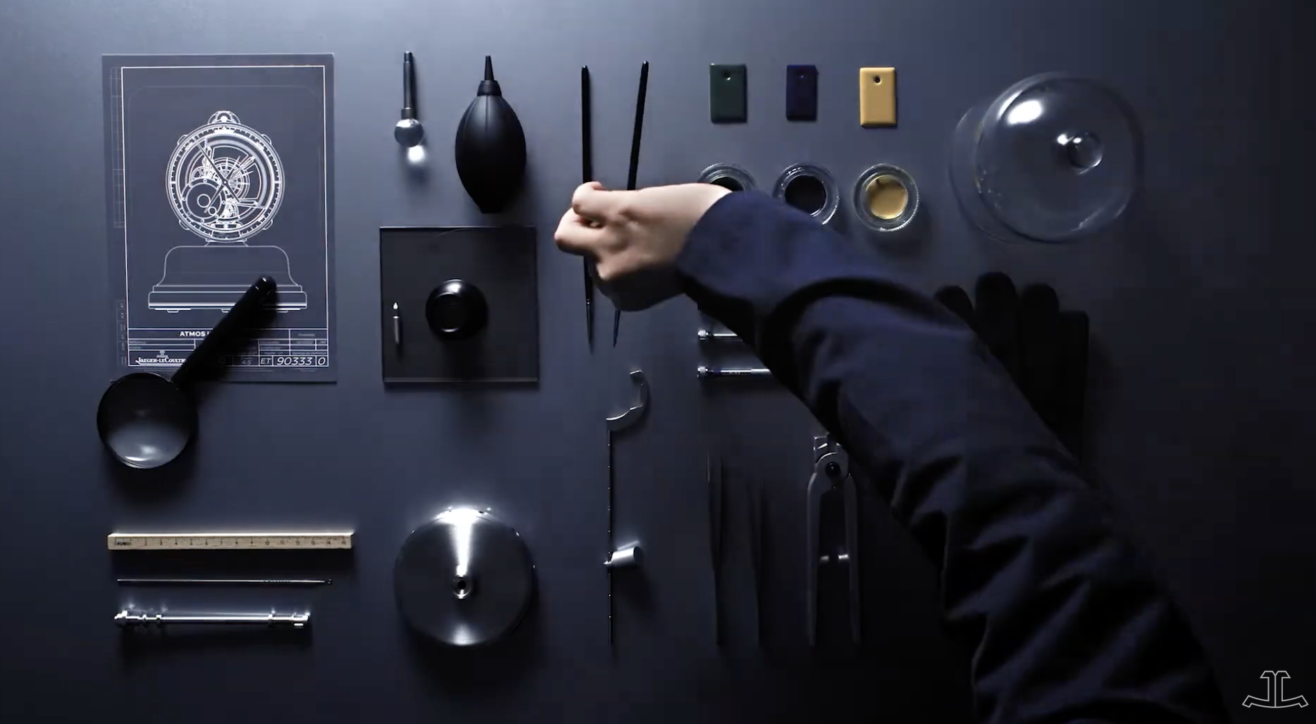 Top-down view of a black table with various watchmaking tools and parts, including a schematic drawing of a watch, a hand holding tweezers, a loupe, small containers of watch components, and metal watch parts, all arranged neatly for assembly or repair.