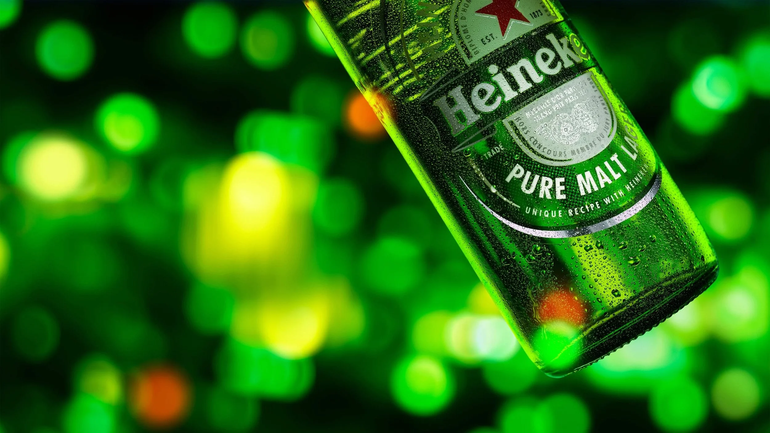 A close-up of a Heineken beer bottle covered in condensation with a blurred background of green and yellow lights.