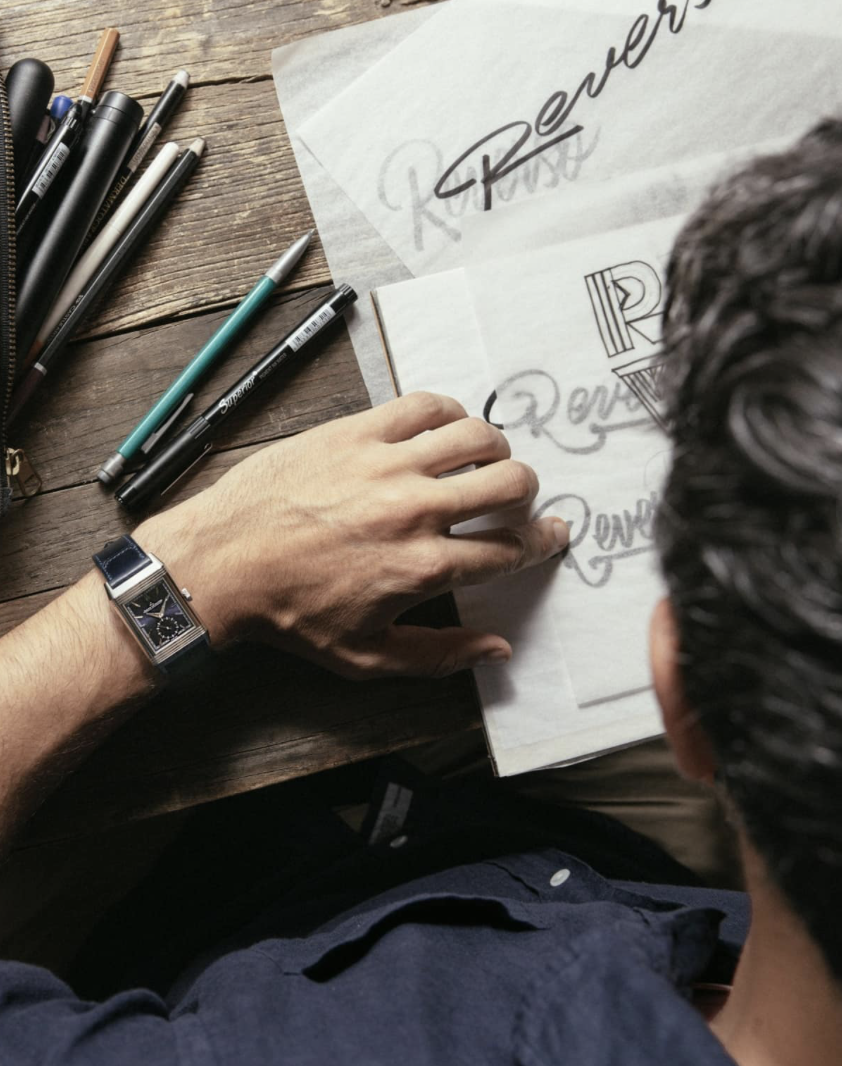 A person designing hand-lettered logos for the word "Review" on paper, surrounded by black and gray pens and markers on a wooden table.