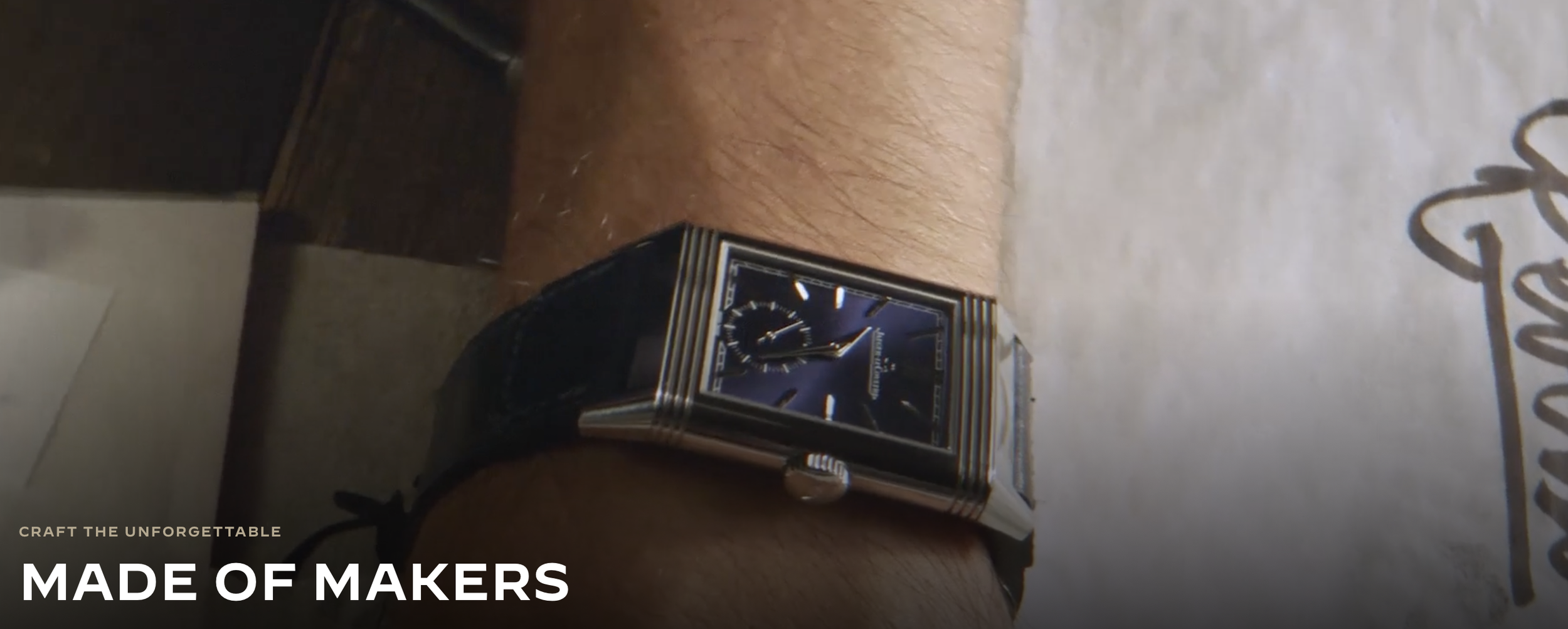 A person's wrist wearing a rectangular silver watch with a black dial, silver hour markers, and a patterned bracelet band, resting on a surface with a white paper or fabric and a brown object in the background.