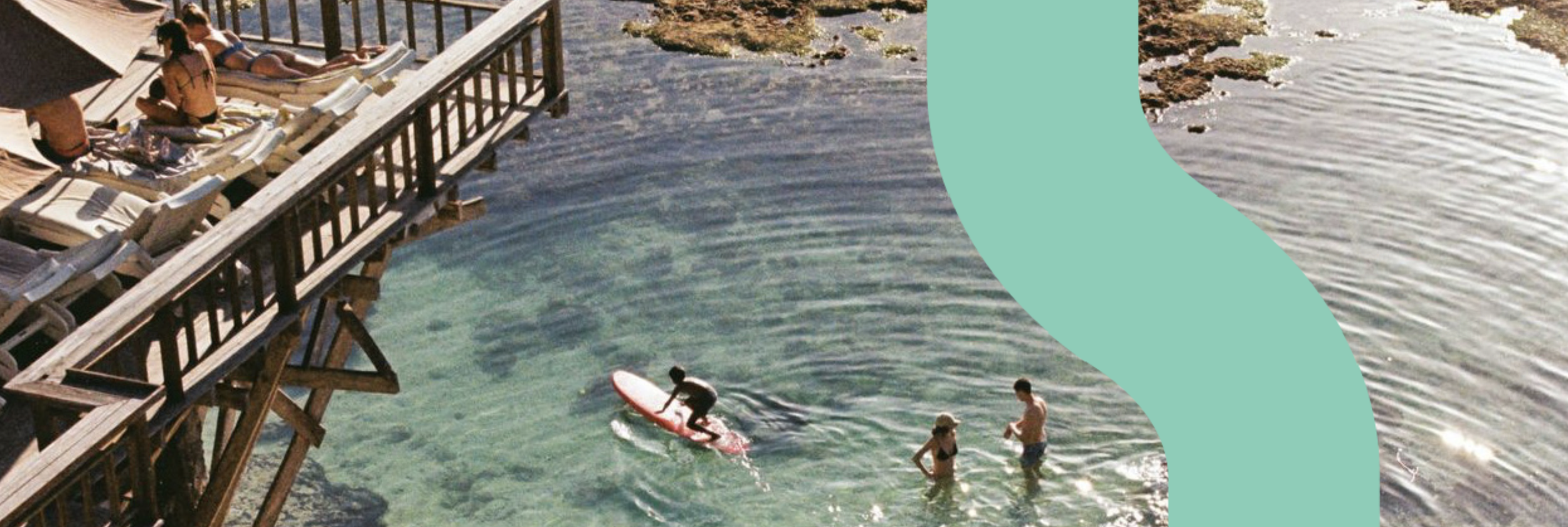 People relaxing on a wooden pier and swimming in clear water near the shore, with some on a paddleboard.