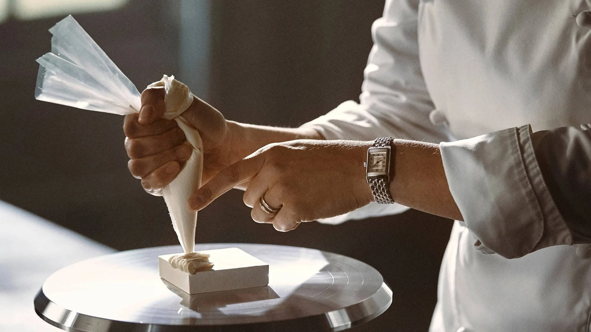 Person in a white coat squeezing cream from a pastry bag onto a small rectangular block on a turntable.
