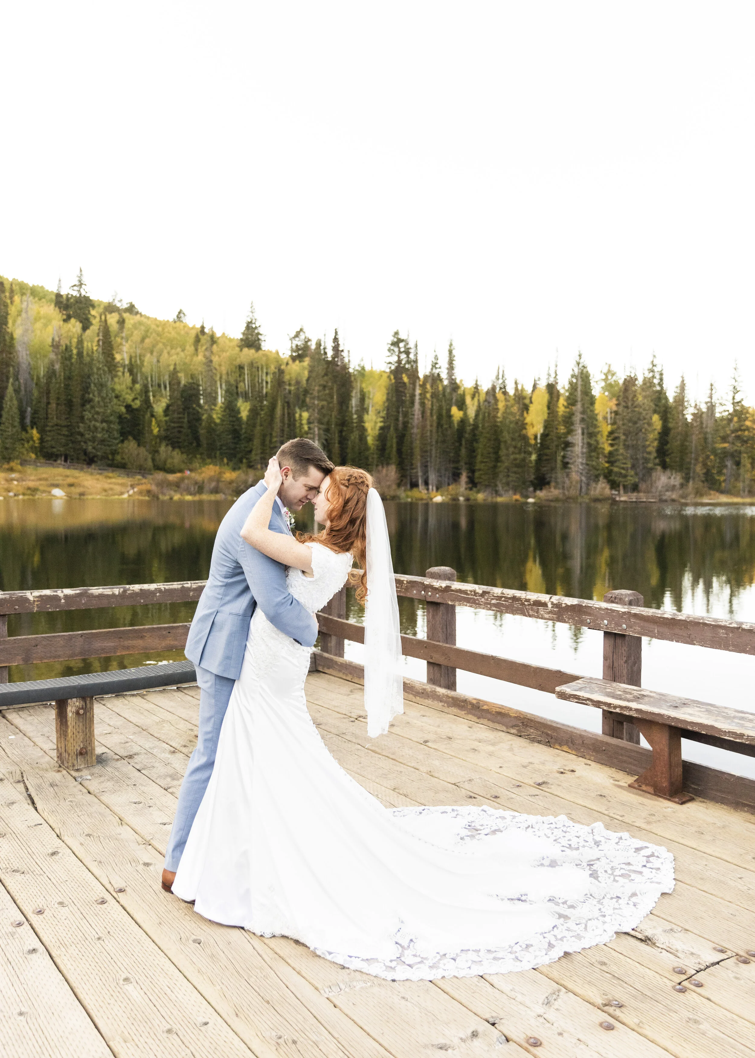  Bride and groom holding each other on a rustic dock lakeside bridals rustic lake wedding photos wooden dock lace train full train wedding gown veil placement inspiration hugging pose wedding day pose ideas forest background capturing nature in weddi