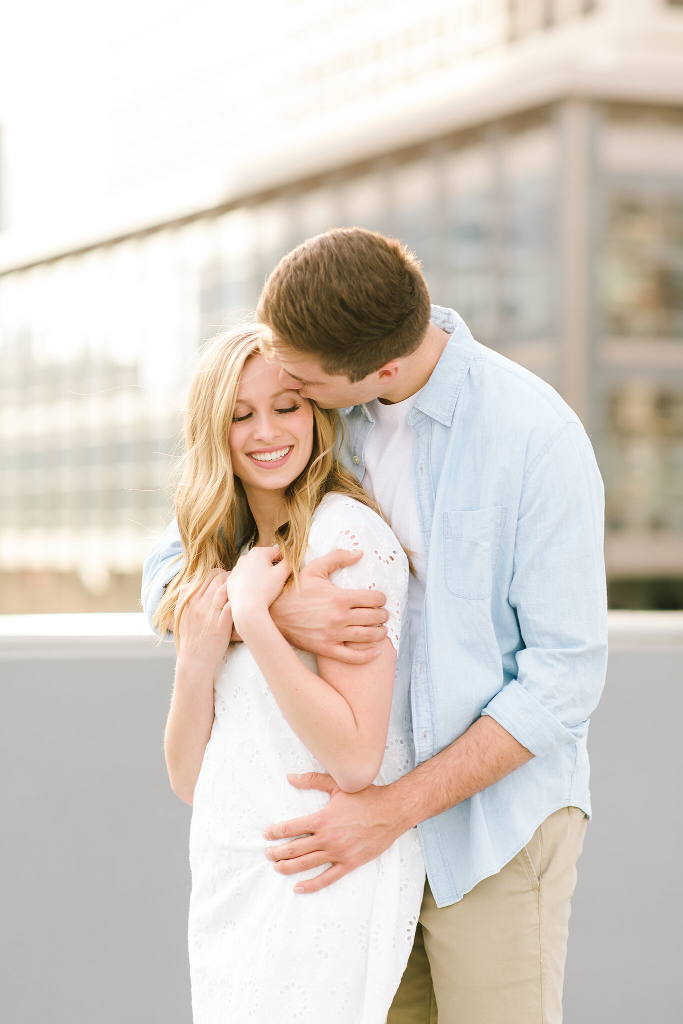  kiss on the forehead arms wrapped around each other engagement session smiling light blue collared shirt white lace short sleeve dress downtown engagement session roof top engagement session salt lake city utah professional utah valley engagement ph