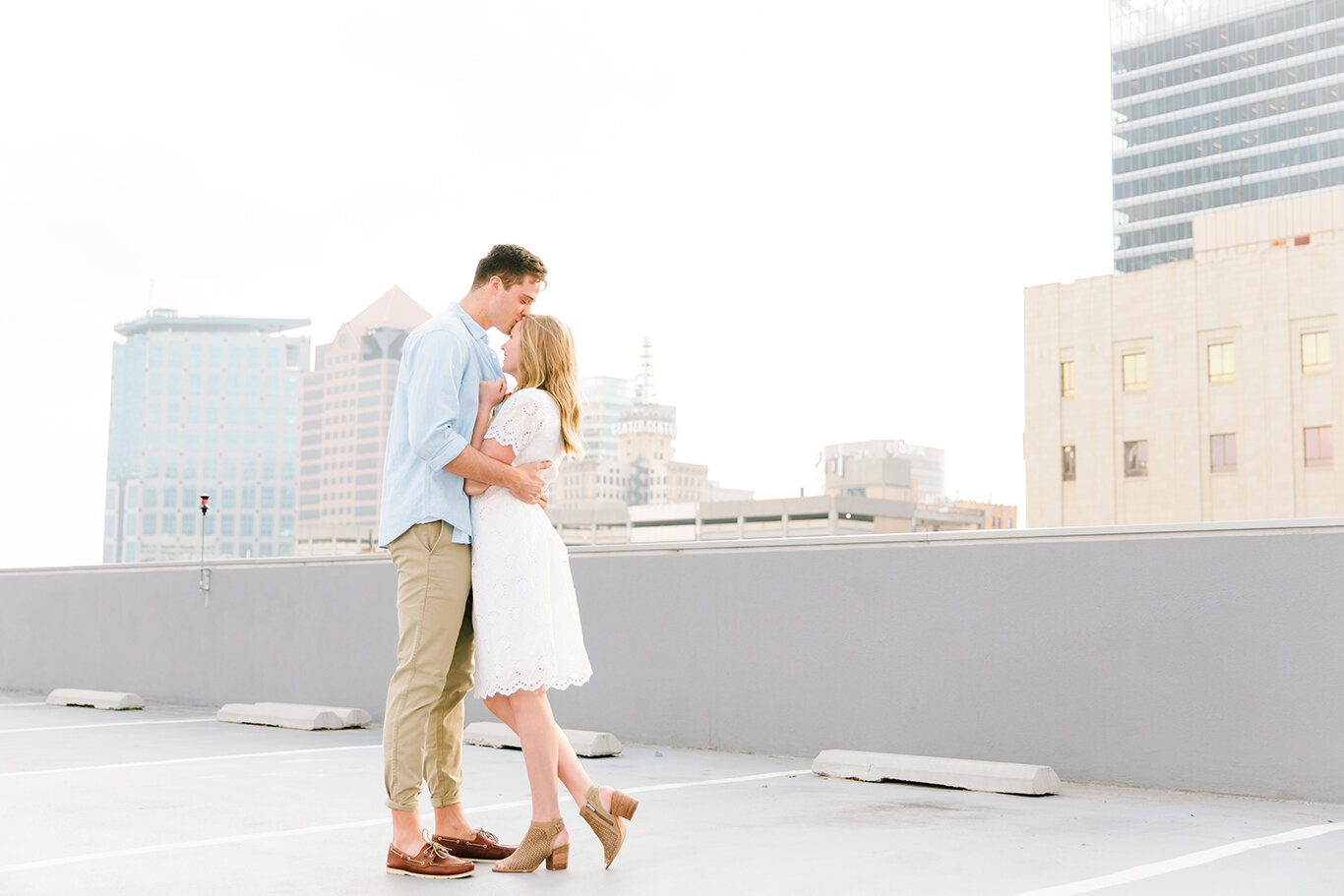  rooftop engagement photo session kiss on the forehead downtown engagement shoot light blue collared casual shirt white lace short sleeve dress loose hanging curls salt lake city utah professional utah valley engagement photographer #downtownsaltlake