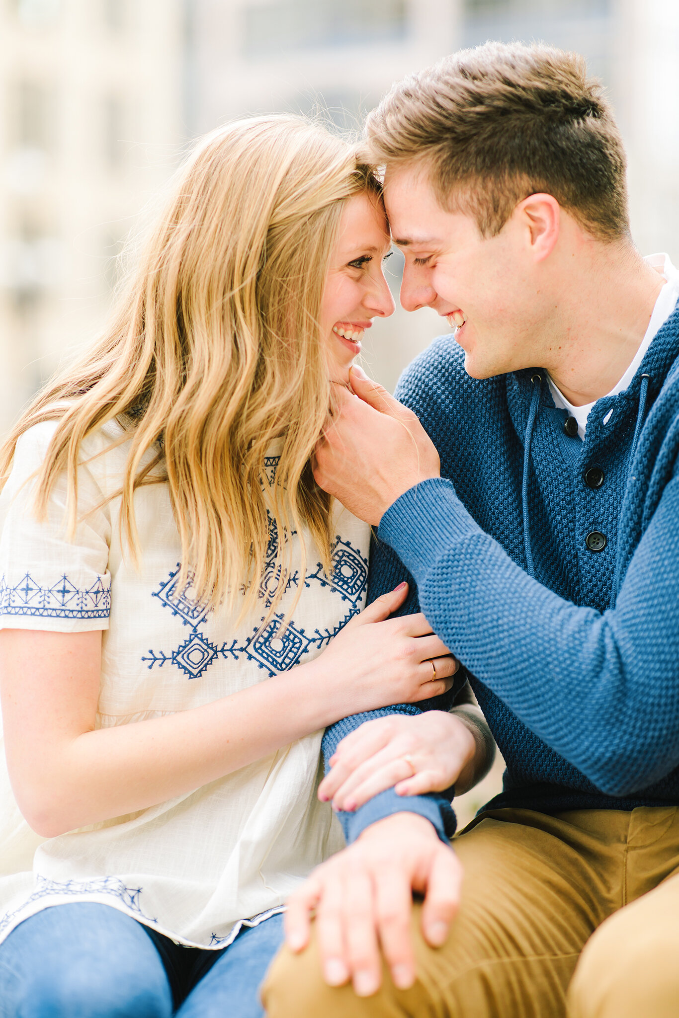  smiling forehead to forehead sitting in downtown salt lake city utah his hand on her chin locked eyes loose hanging curls navy blue sweater  navy blue thread detailed shirt salt lake city utah professional utah valley engagement photographer #downto