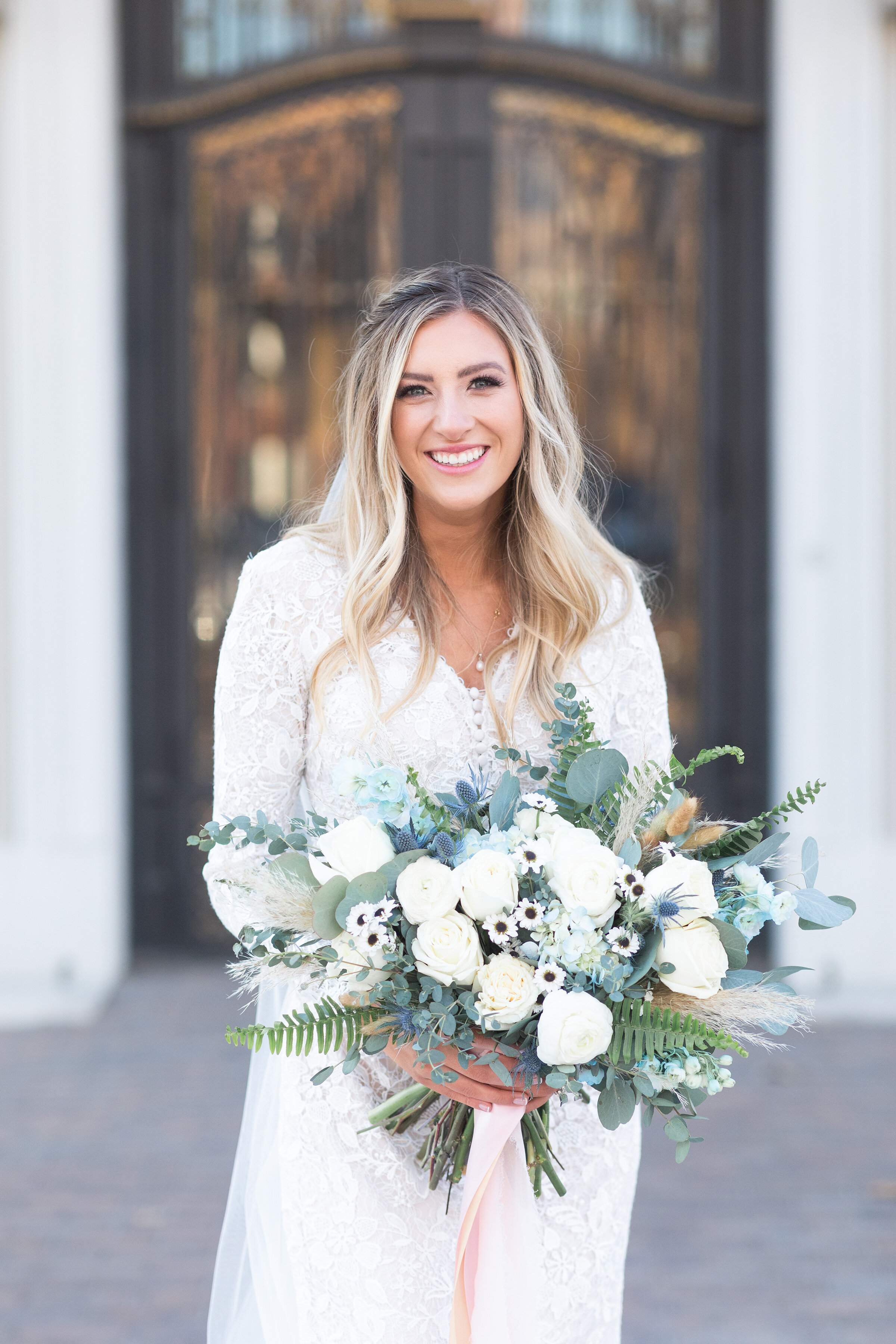  Salt Lake City’s Clarity Lane Photography captures a happy, smiling bride pose outside of the Monson Center building. long blonde loose waves balyage wedding hair, blue and white bohemian wedding bouquet, long sleeve lace wedding dress, monson cente