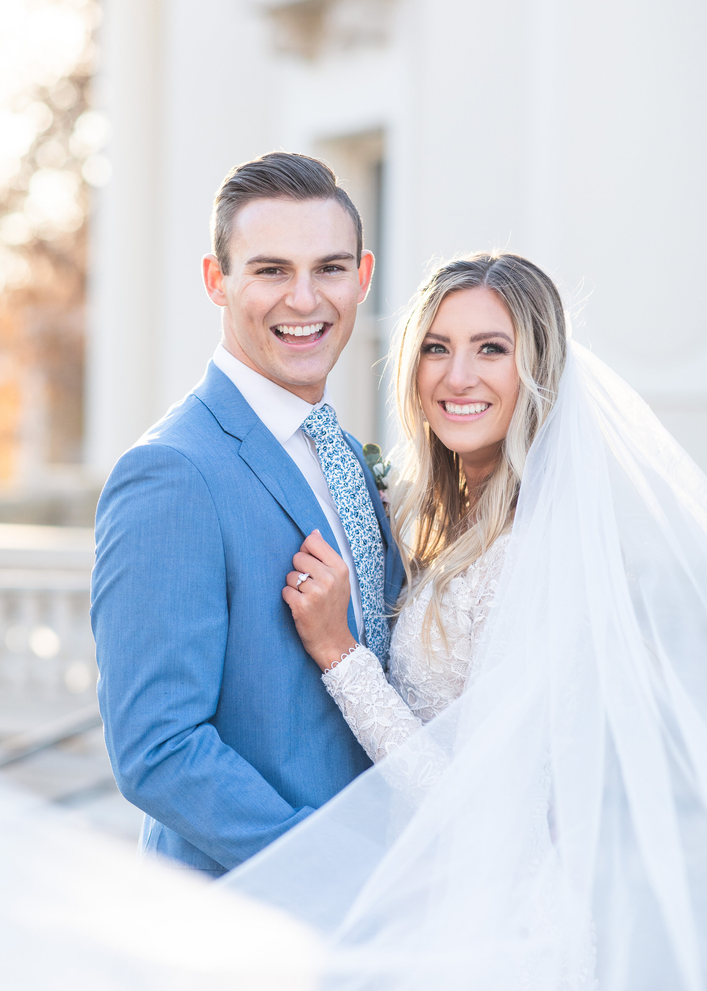  Clarity Lane Photography photographs a happy soon-to-be bride and groom outside their Monson Center in Salt Lake CIty, Utah. long bridal veil train billows in the wind, smiling bride and groom, brides hand on groom chest, stone pillar building, prof