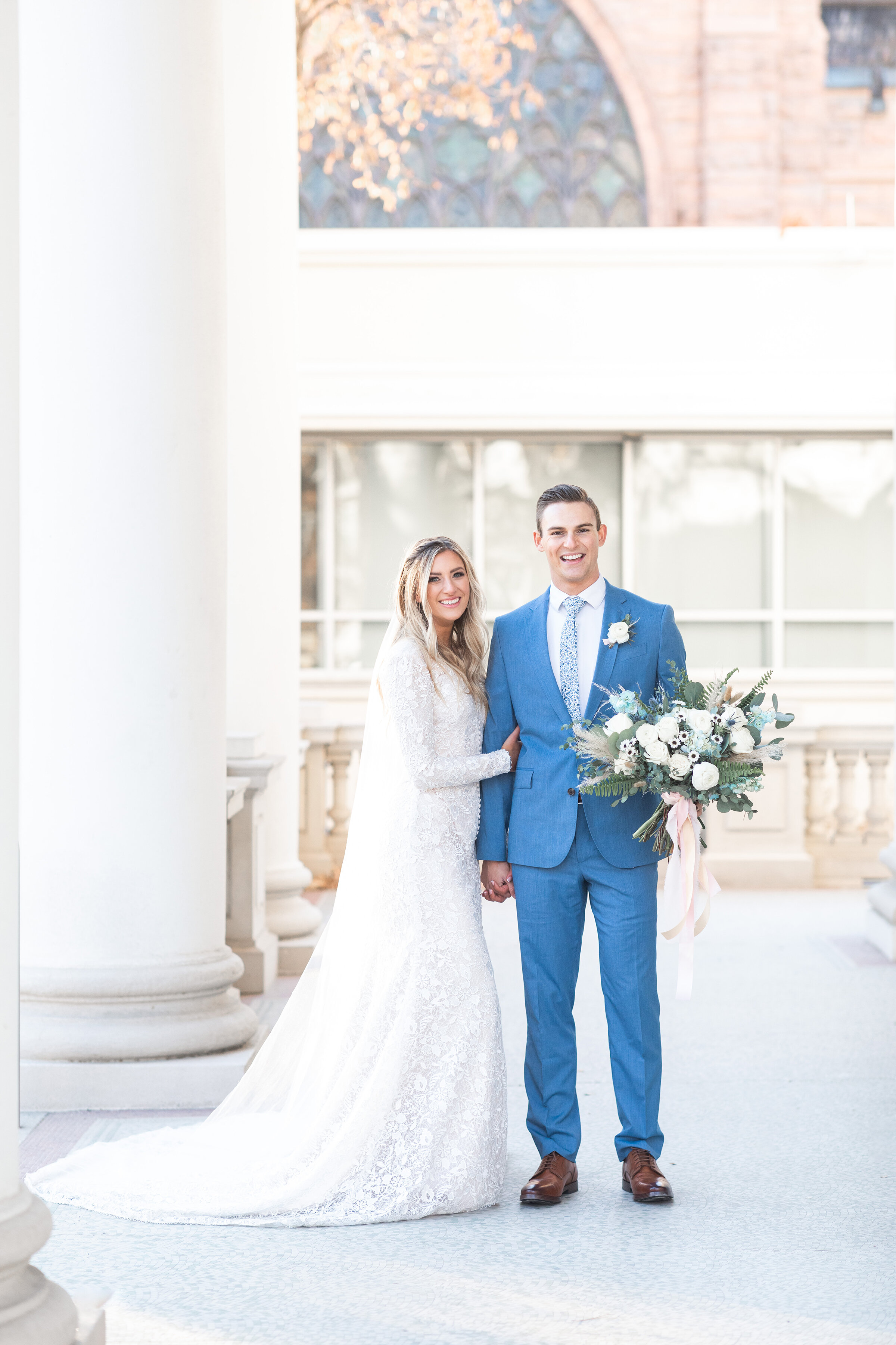  Posed outside the Monson Center in Salt Lake City, Utah, Clarity Lane Photography captures bride and groom arm in arm and smiling. medium blue wedding suit, blue and white wedding colors, blue and white wedding bouquet, long sleeved fitted beaded la