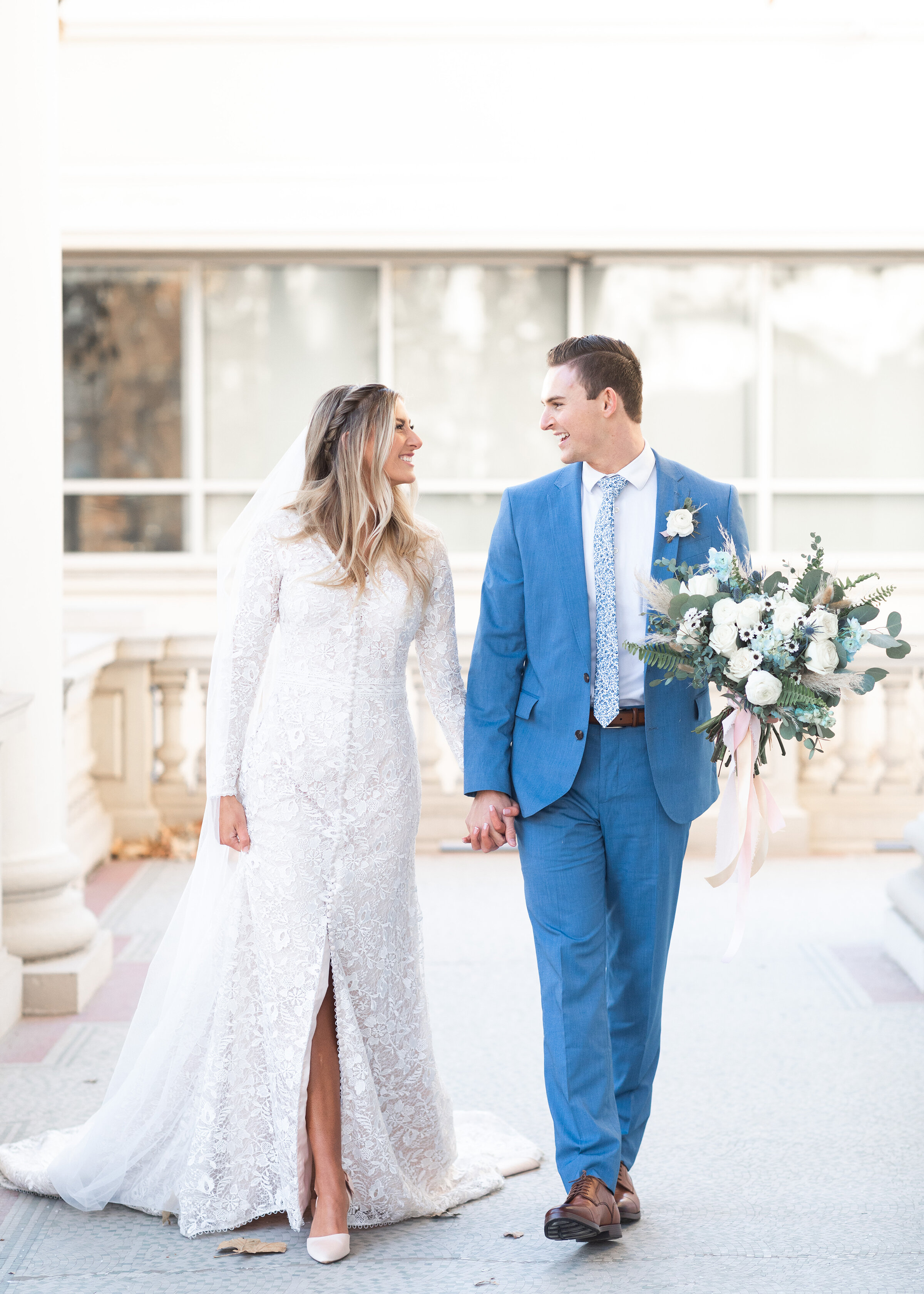  Clarity Lane Photography captures bride and groom hand-in-hand strolling outside the Monson Center in Salt Lake City, Utah. long sleeved beaded lace wedding dress, front slit in wedding dress, long blonde balayage half up bridal hair, long bridal ve