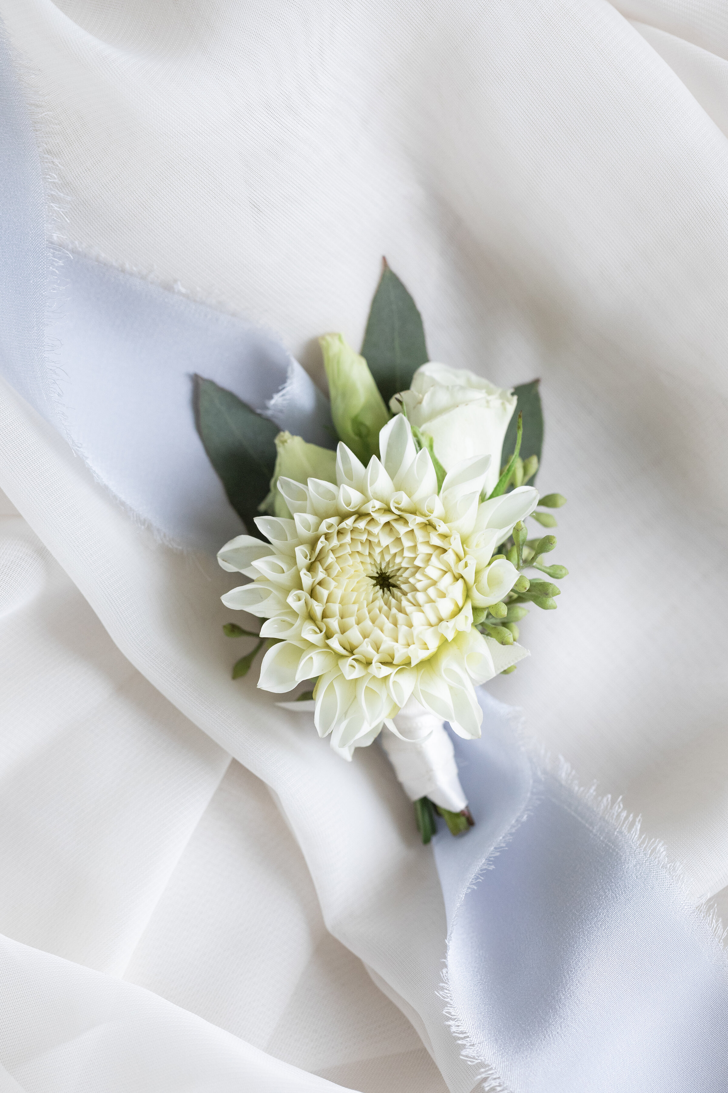  Pressed against a soft chiffon background, Clarity Lane Photography captures wedding day details including this white boutonnière. white daisy boutonniere, white wedding flowers, wedding flower flatlay photo, white chiffon flatlay background, profes