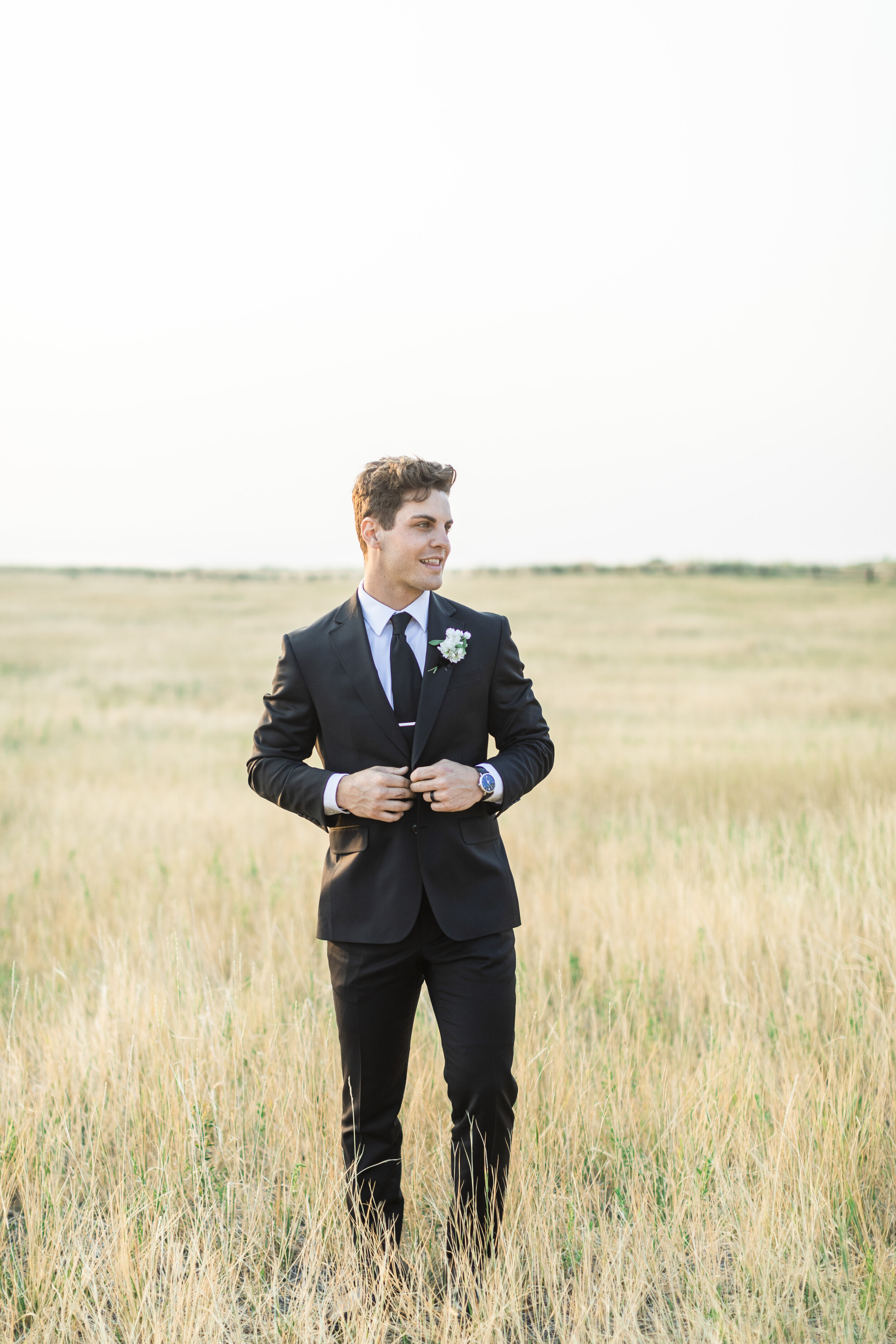  Groom wearing a black suit with a black tie stands in a field at tunnel springs and buttons his suit coat captured by high-end wedding photographer Savanna Richardson Photography. Groom buttons suit slc weddings groom style groom fashion #savannaric