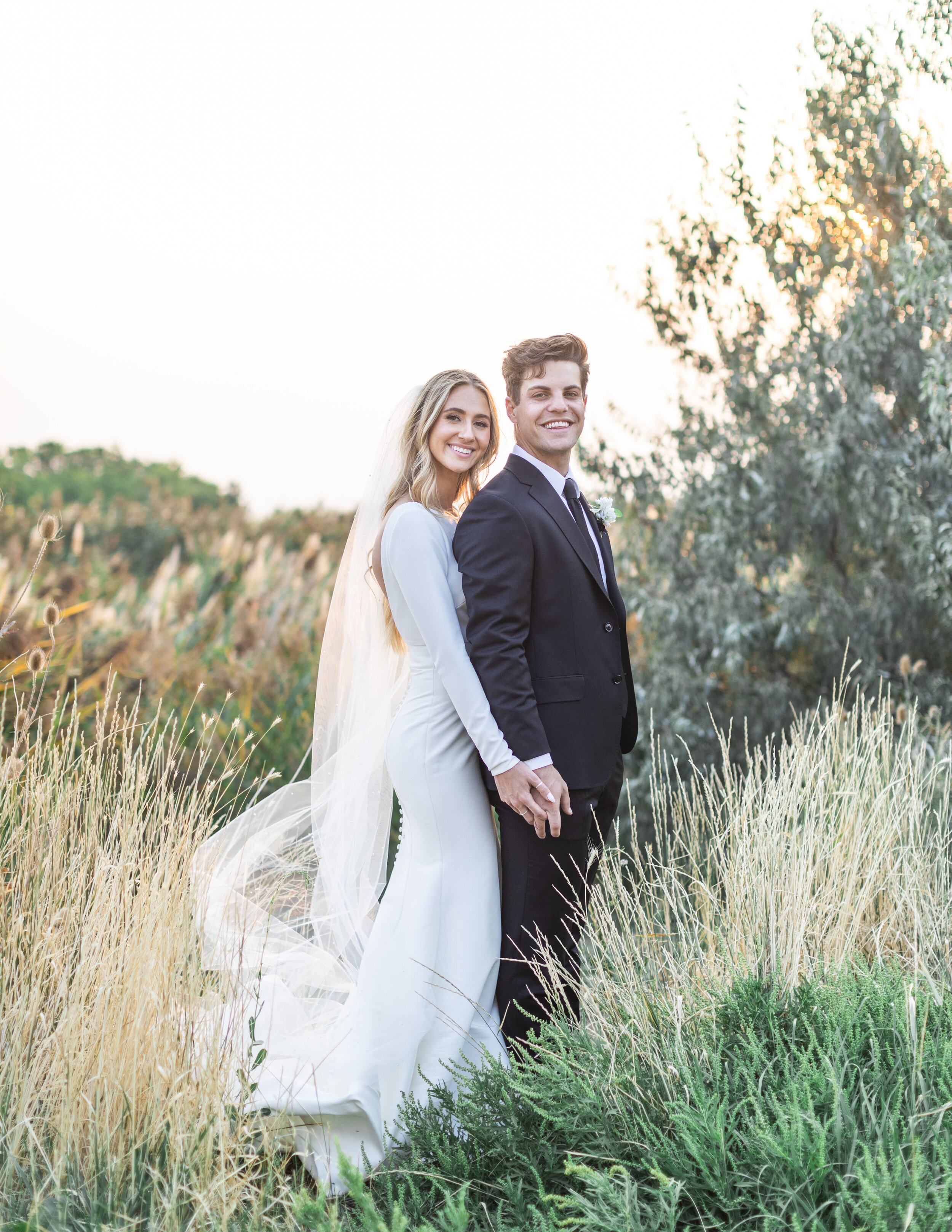  A Salt Lake City photographer captures a bride and groom holding hands and smiling at sunset in a yellow grass field with the bride's veil blowing in the wind captured by Savanna Richardson Photography. Golden sunset backlighting on photos dreamy ut