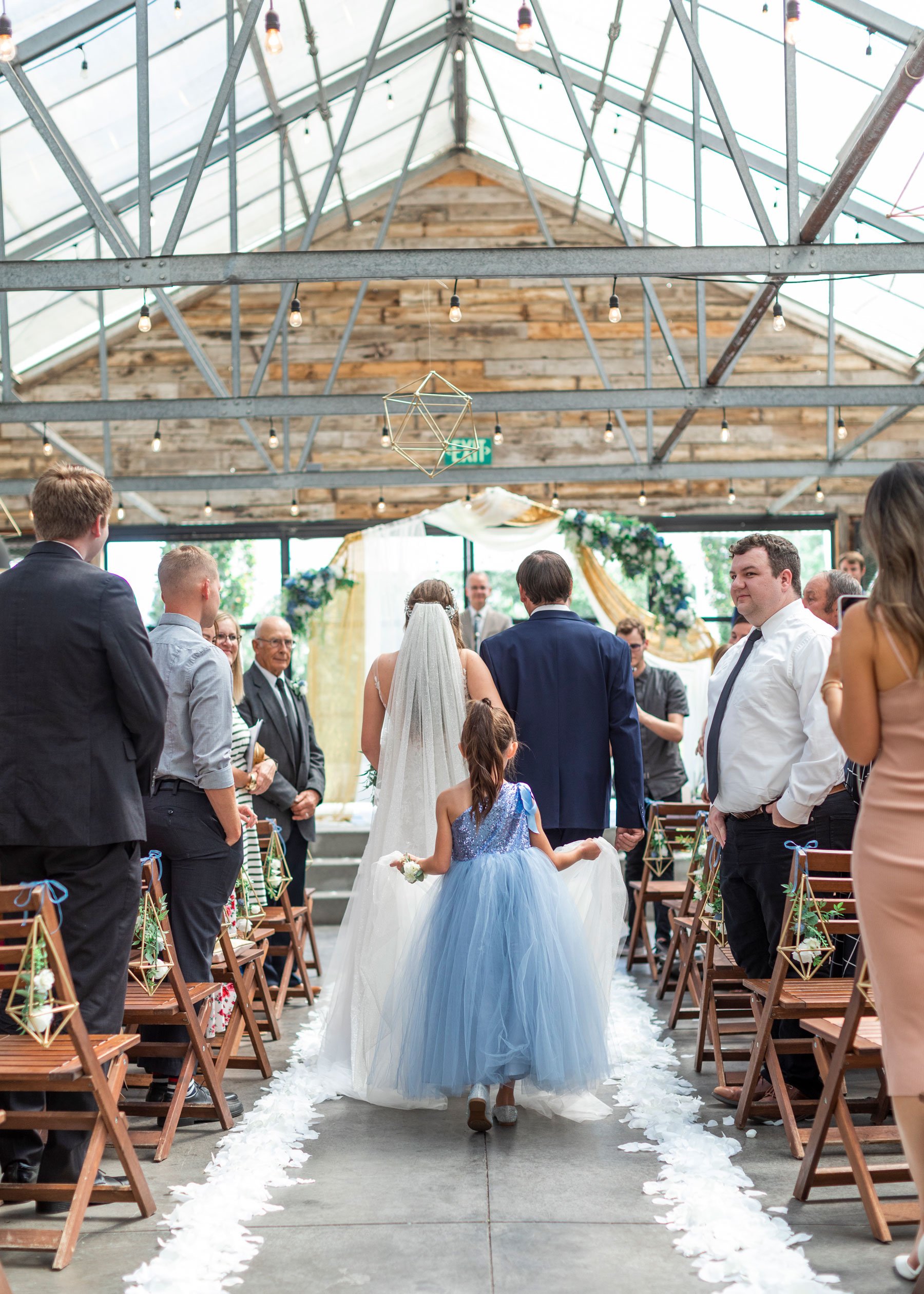  A professional wedding photographer Savanna Richardson Photography captures a darling picture of the bride walking down the aisle with a girl holding her wedding train behind her. Holding the wedding dress little girl walks down the aisle here comes