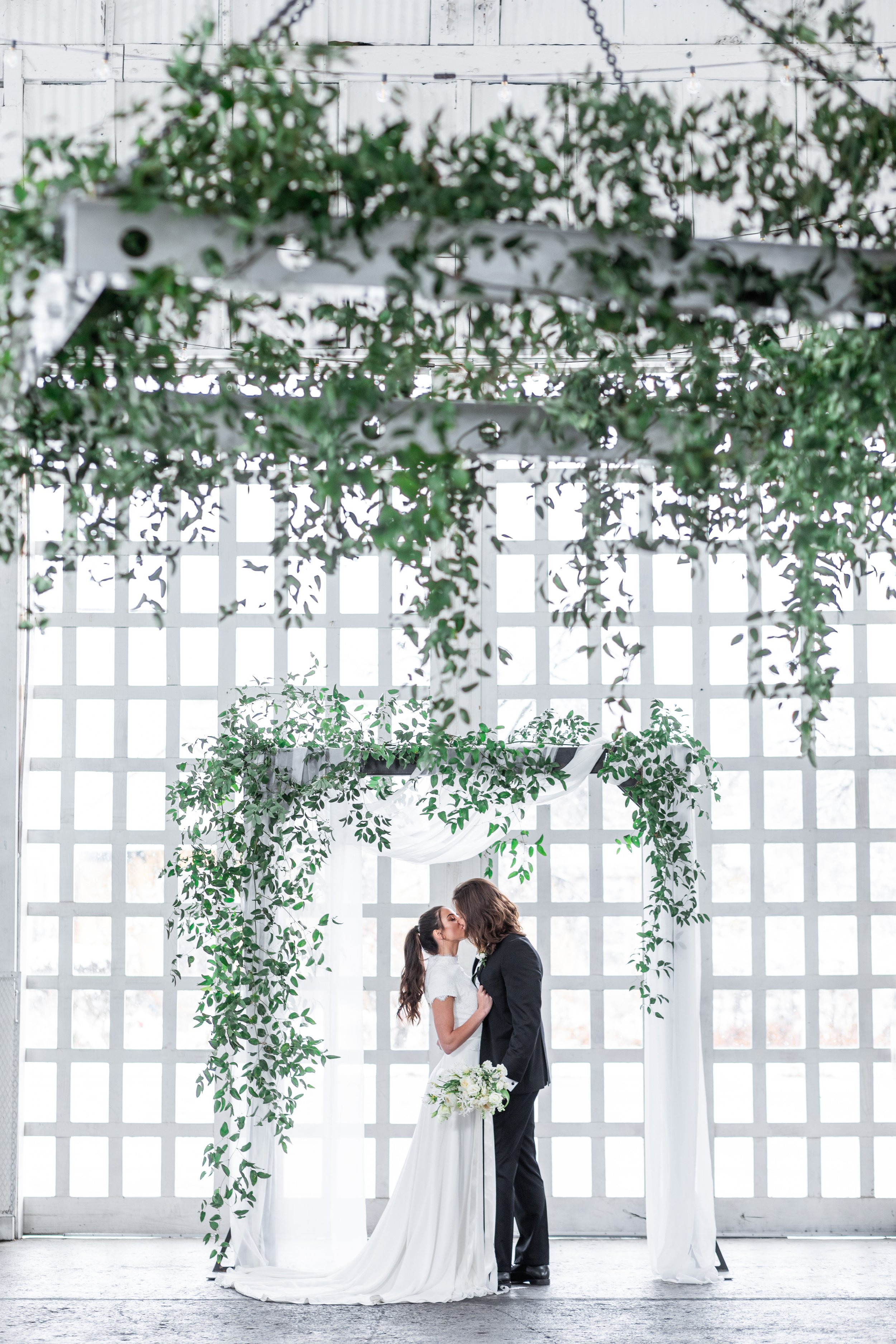  A groom and bride kiss in front of a wall of windows and fresh greenery by Savanna Richardson Photography. stunning altar ideas wall of windows #savannarichardsonphotography #utahvendor #utahweddingphotographer #MilleFleurDesign #Utahweddingdesigns 