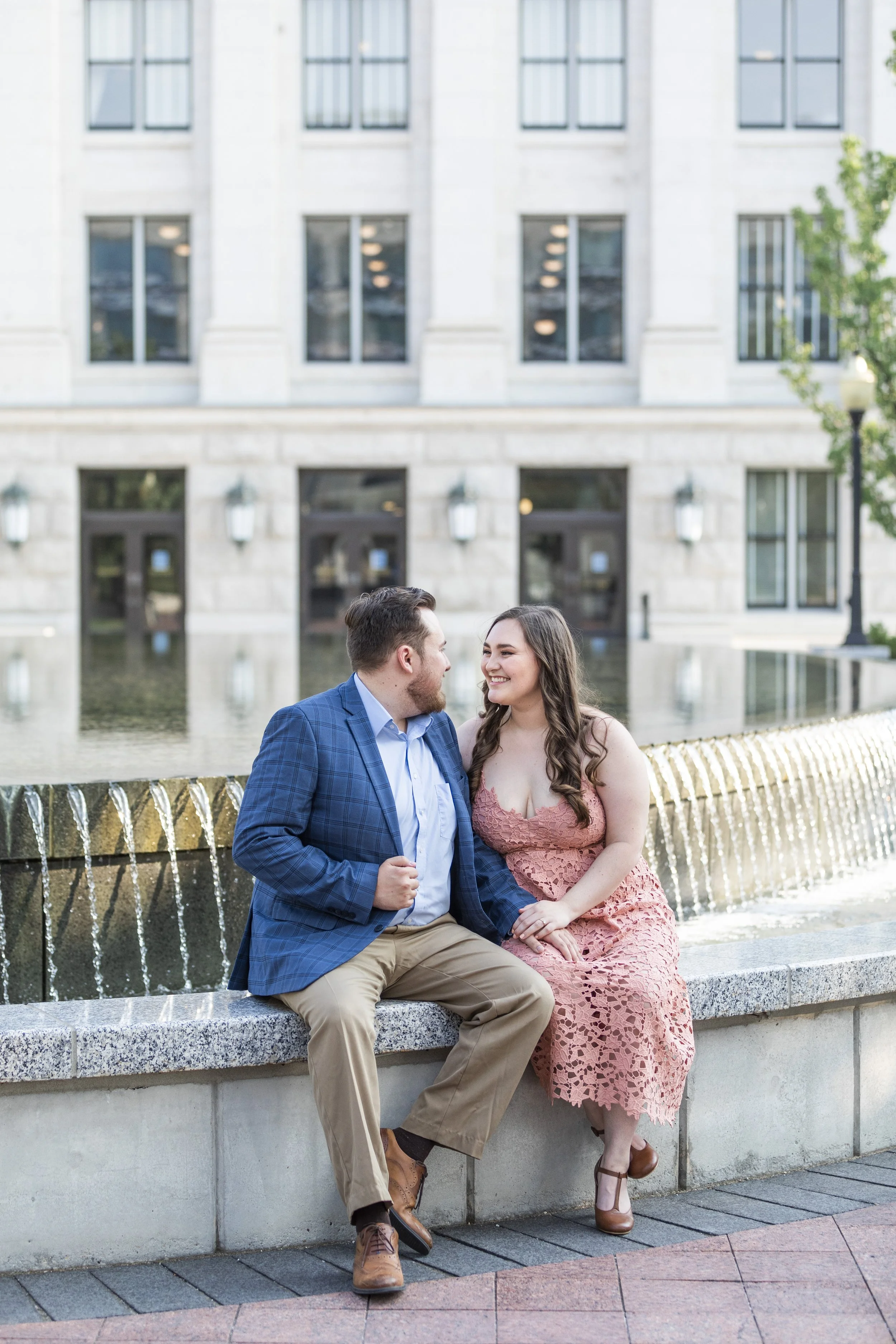  In front of a water fountain, a couple sits together during a Utah engagement session by Savanna Richardson Photography. Utah professional photographer #savannarichardsonphotography #Utahcapitolphotography #utahengagements #utahengagementphotographe