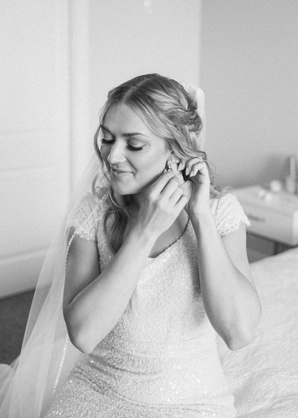  Black and white photo of a blonde bride in a sparkly wedding gown puts on her earrings before the wedding ceremony. #bridalinspo #summerbride #shortsleeveweddingdress #modestweddingdress #savannarichardsonphotography #cachevalley #gettingready 