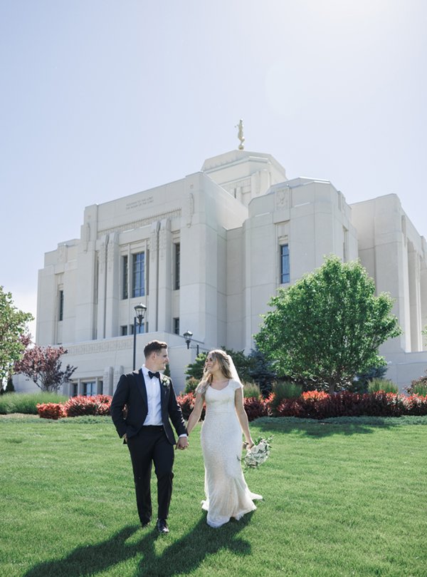  The happy couple walking hand in hand across the lawn in front of the LDS temple in Meridian, Idaho on a summer day. #ldstemplewedding #brideandgroom #groomfashion #modestbridalfashion #cachevalley #utahweddingphotographer #summerwedding #tonygrove 