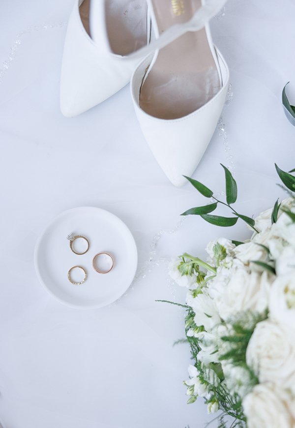  Detail shot of a bride’s white pointed heels next to the wedding rings and her cream wedding bouquet on a white marble background. #savannarichardsonphotography #weddingdetails #ringinspo #utahweddingphotographer #cachevalley #summerwedding #bride 