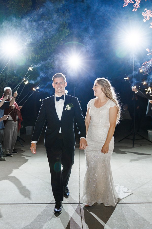  Bride and groom exit their wedding with friends and family waving sparklers along the walkway and cheering for the couple. #savannarichardsonphotography #weddingexits #sparklerexit #outdoorweddingreception #summerwedding #cachevalley #tonygrove 