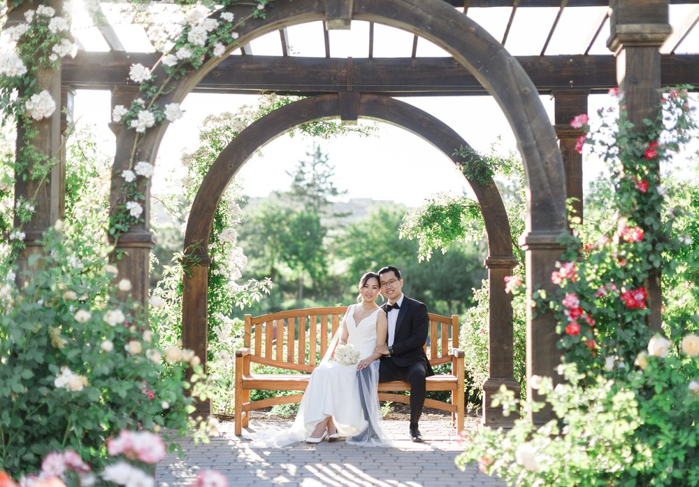  Bride and groom sitting close on a bench under a pergola surrounded by flowers in Ashton Gardens. #savannarichardsonphotography #thanksgivingpoint #utahweddingphotographer #groomfashion #formalposeinspo #weddingdress #summerwedding #coupleposeideas 