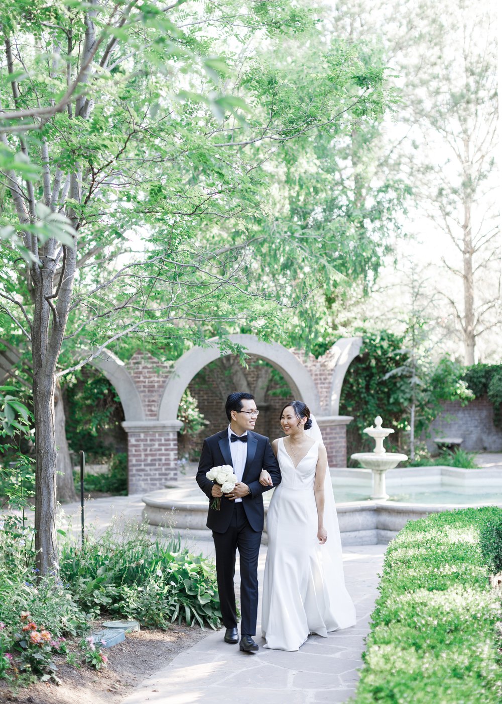  Happy couple walking down the walkway at the fountain at the Thanksgiving Point Ashton Gardens. #savannarichardsonphotography #thanksgivingpoint #utahweddingphotographer #groomfashion #formalposeinspo #weddingdress #summerwedding #coupleposeideas 