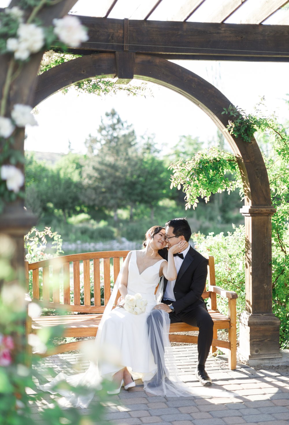  The happy couple sitting on a bench sharing a kiss while surrounded by flowers in Ashton Gardens. #savannarichardsonphotography #thanksgivingpoint #utahweddingphotographer #groomfashion #formalposeinspo #weddingdress #summerwedding #coupleposeideas 