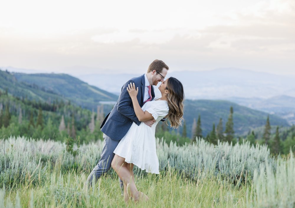  In a grass meadow, a man leans over and kisses his fiance in Park City Utah by Savanna Richardson Photography. mountain overlook dusk #ParkCityEngagements #ParkCityPhotographers #SavannaRichardsonPhotography #SavannaRichardsonEngagements 