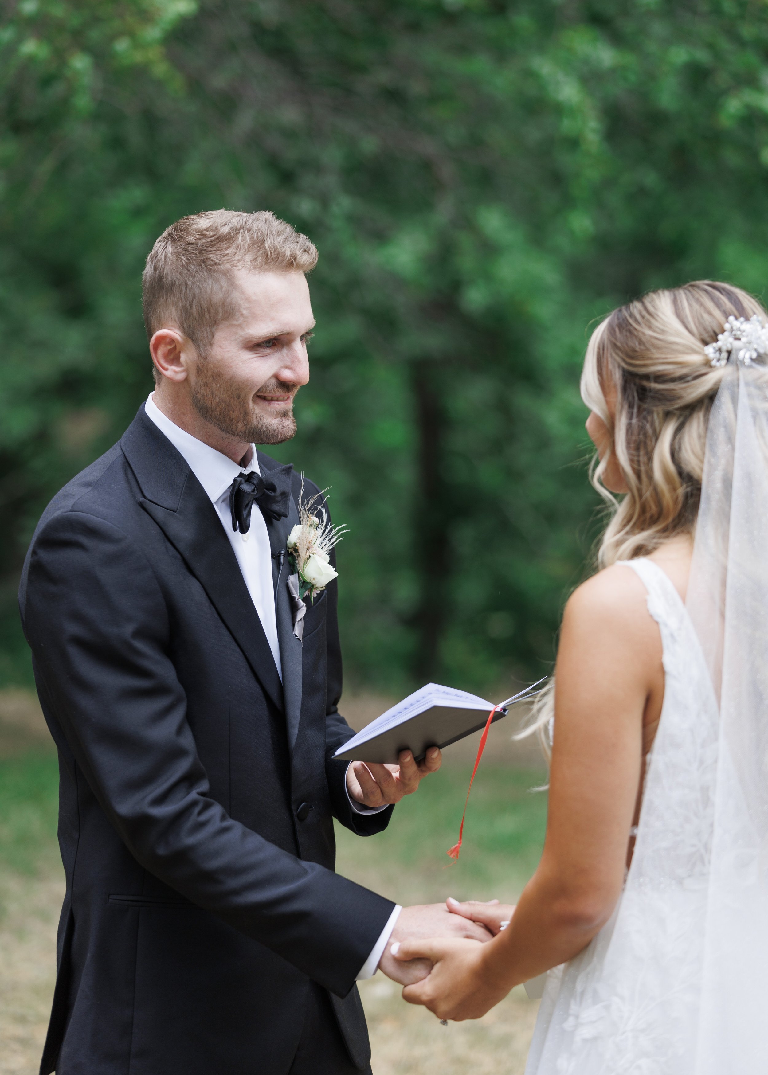  A groom in a black suit and bowtie holds the bride’s hands while reading his vows by Savanna Richardson Photography. groom vows Mapleton #SavannaRichardsonPhotography #SavannaRichardsonWeddings #QuietMeadowFarms #MapletonUTweddingphotographers 