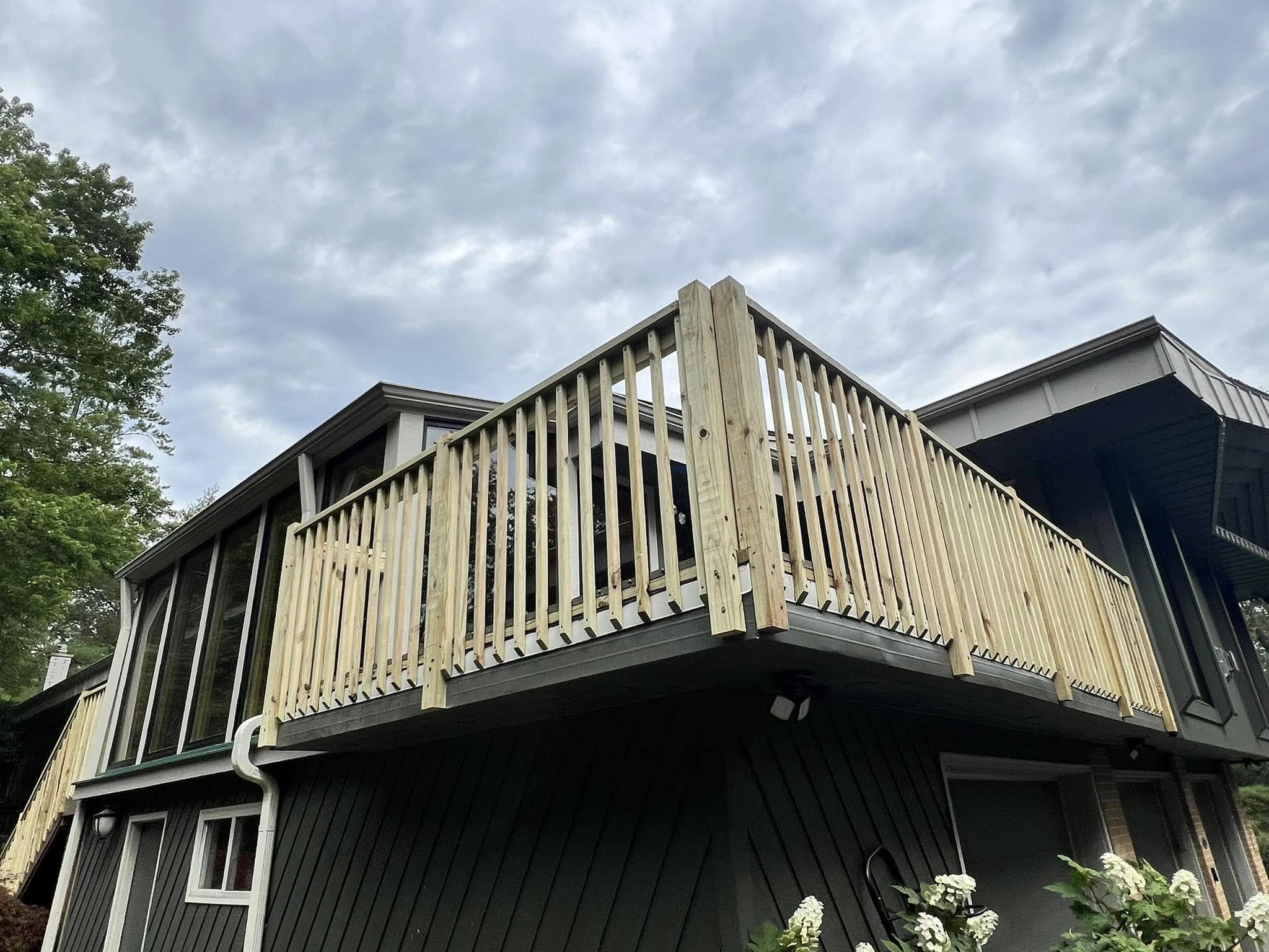 The image shows the upper part of a house with a balcony made of wood and a black sliding door. There's a tree on the left side and a cloudy sky overhead.