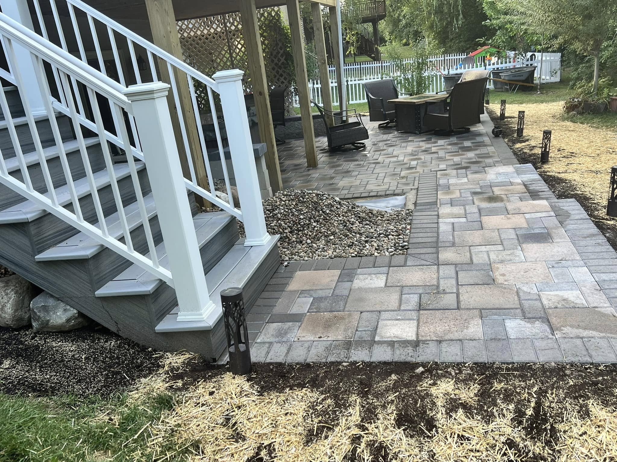 Backyard patio area with a brick paved floor, outdoor furniture including a table and chairs, surrounded by a white picket fence and trees.
